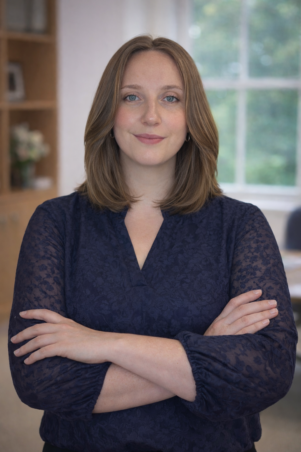 Close-up of a young woman with long, light brown hair and blue eyes, smiling gently, wearing a black turtleneck, brown blazer, and gold earrings.