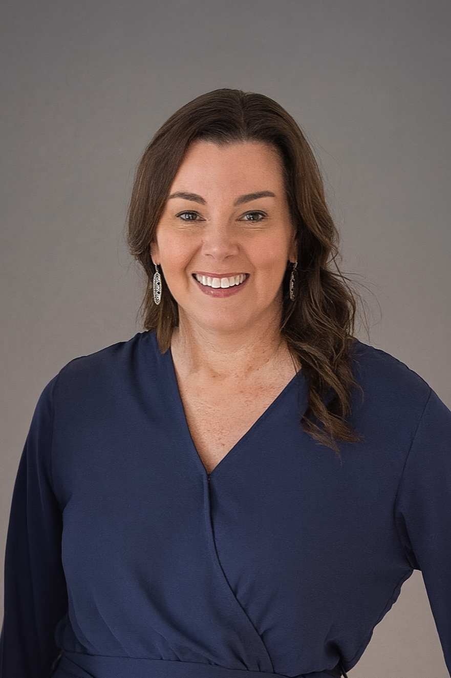 A woman with brown hair smiling, wearing a navy blue blouse and silver earrings, posed against a gray background.