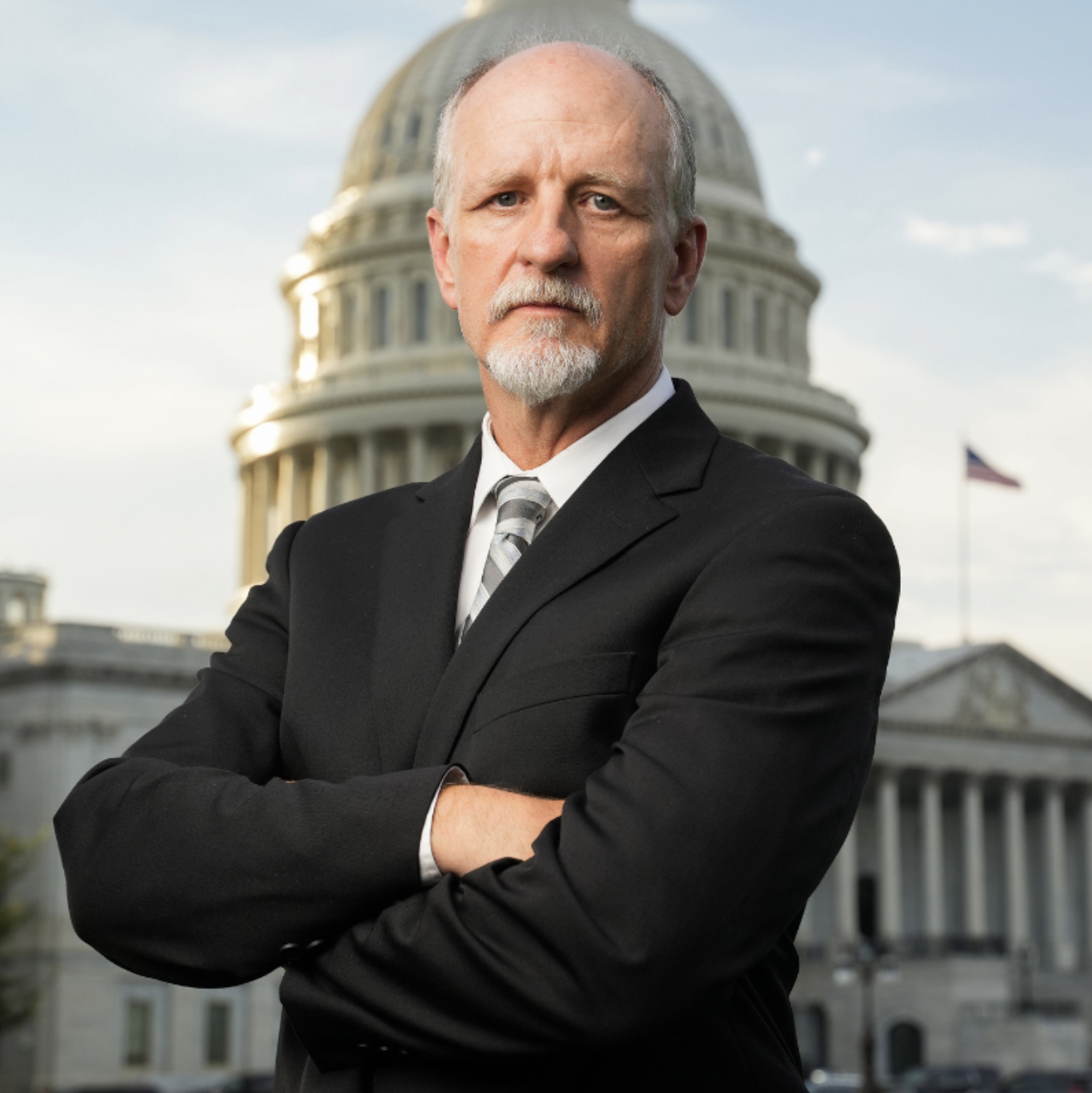 A stern-looking man with gray hair and a beard, wearing a black suit and striped tie, standing with crossed arms in front of the U.S. Capitol building.
