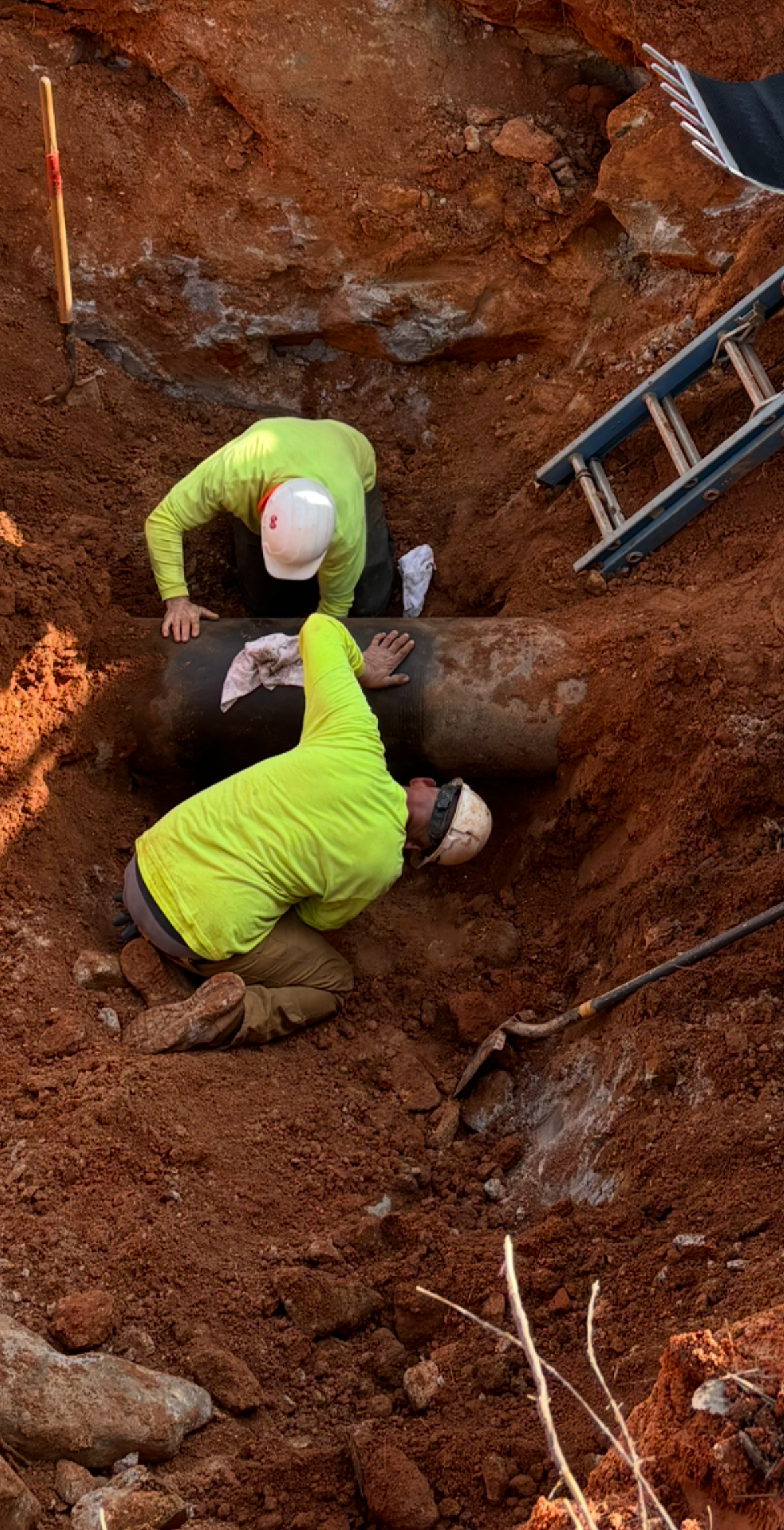 Two construction workers in hard hats and neon yellow shirts installing a large pipe underground, surrounded by reddish dirt with tools and equipment nearby.