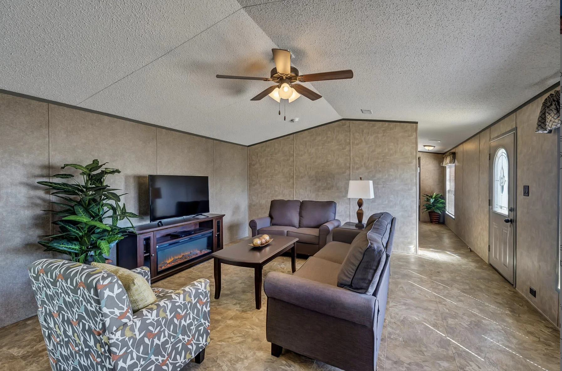Living room with beige walls, a ceiling fan, a flat-screen TV on a wooden stand, a gray loveseat, a brown armchair, a floor lamp, and a small coffee table, with a door and windows in the background.