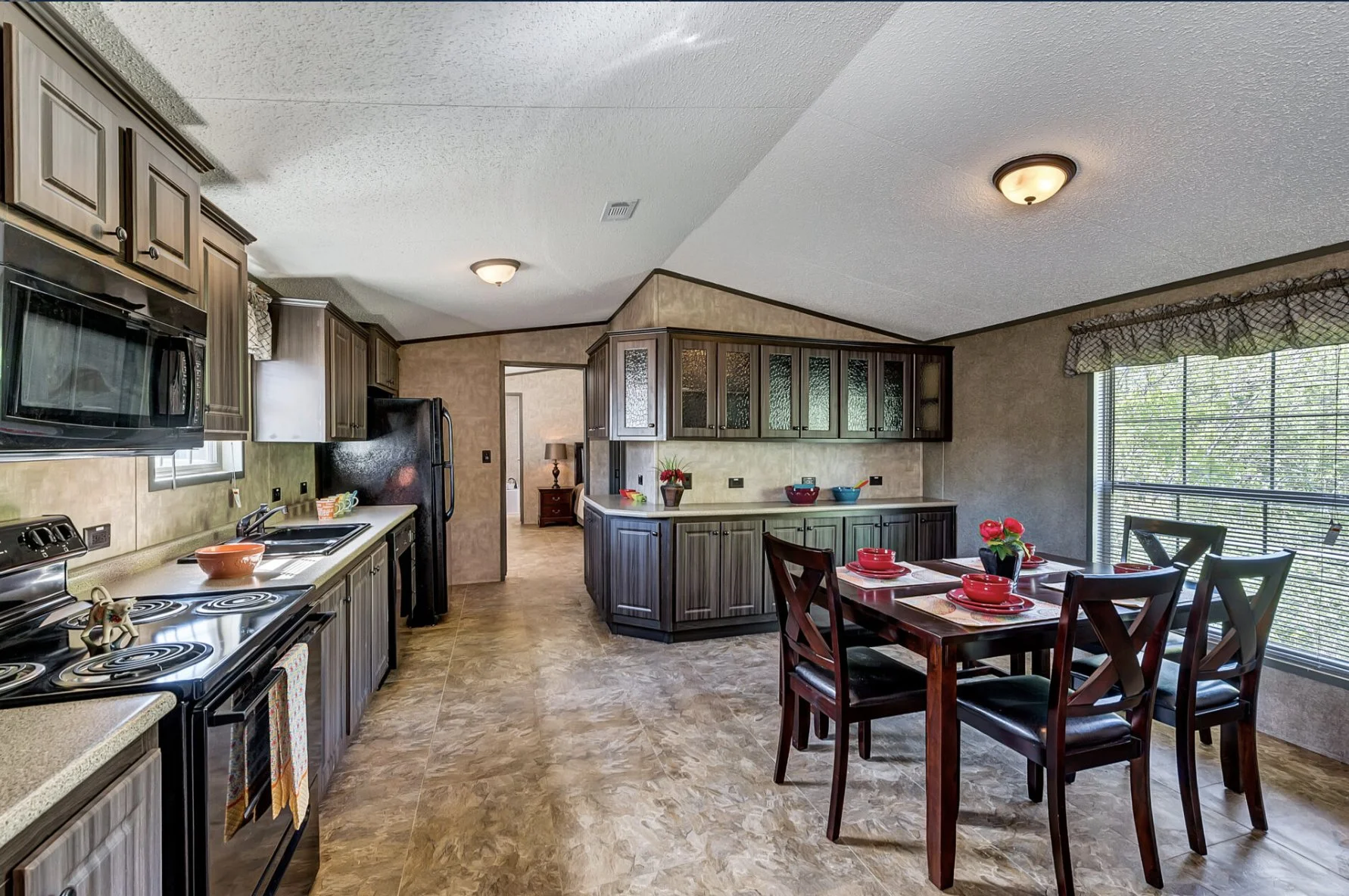 Kitchen and dining area with dark wood cabinets, beige countertops, black appliances, and a wooden dining table set for four with red dishes, next to a large window with blinds and valance.