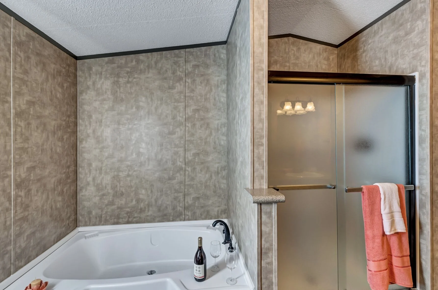 Bathroom corner with a bathtub, glasses, a bottle of wine, and a frosted glass shower door with a towel hanging on a bar.