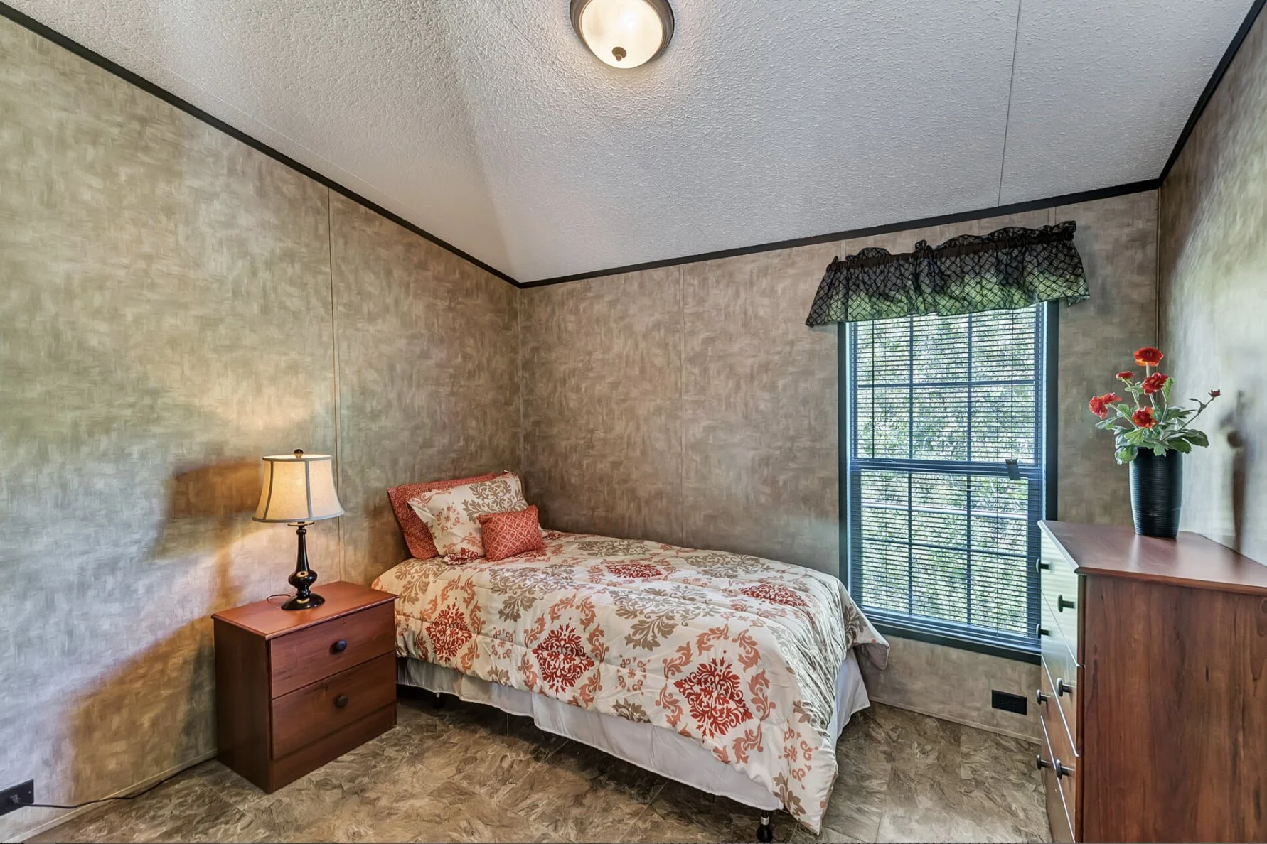 A small bedroom with beige textured walls, a twin bed with a patterned quilt and red pillows, a wooden nightstand with a lamp, a wooden dresser with a vase of red flowers, and a window with blinds and a floral valance.