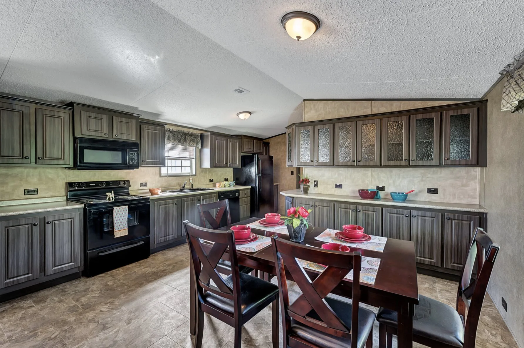 A kitchen with dark wood cabinets, black appliances, a dining table with five chairs, and decorated with bowls, a vase with pink flowers, and placemats.