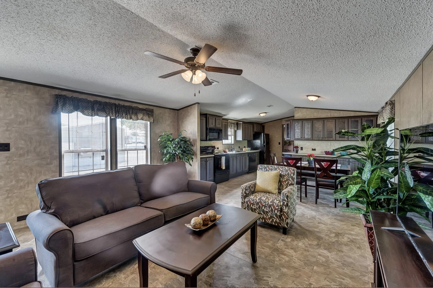 Living room and kitchen area with brown sofa, armchair, wooden coffee table, dining table, and kitchen cabinets with plants and window blinds