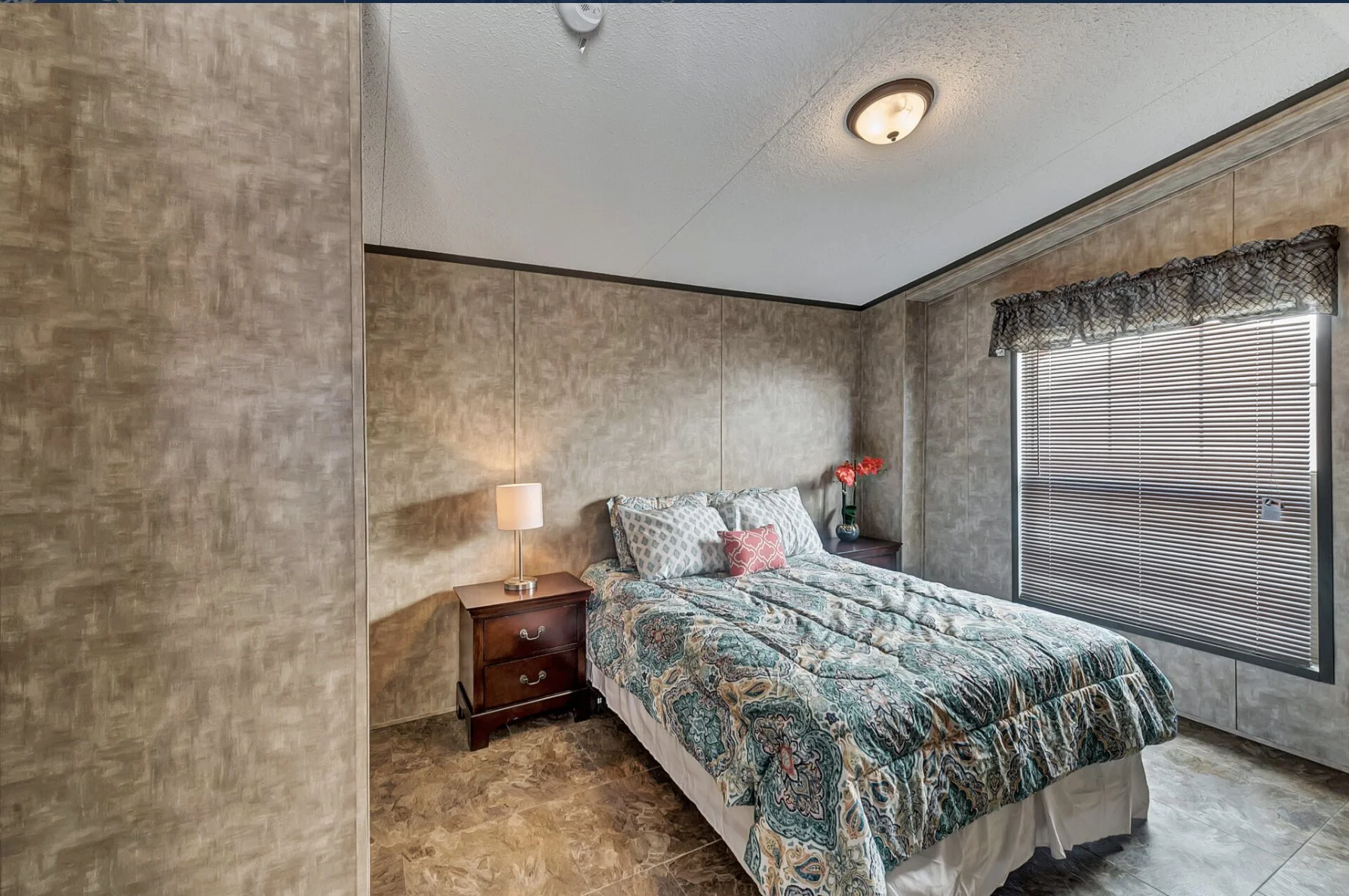 A bedroom with beige textured walls, a bed with a patterned bedspread, and two small wooden nightstands with lamps, one with a vase of red flowers. There is a window with blinds and a valance, and a ceiling light