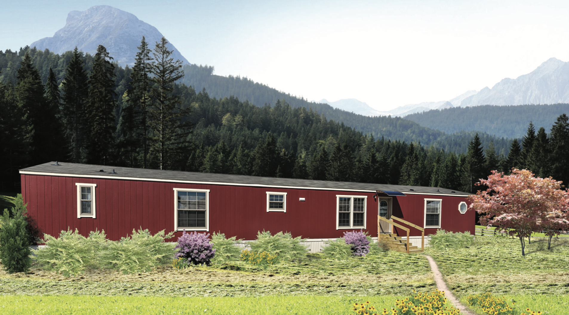 Red manufactured home with a black roof, set in a green field with shrubs and trees, mountains and pine forest in the background.