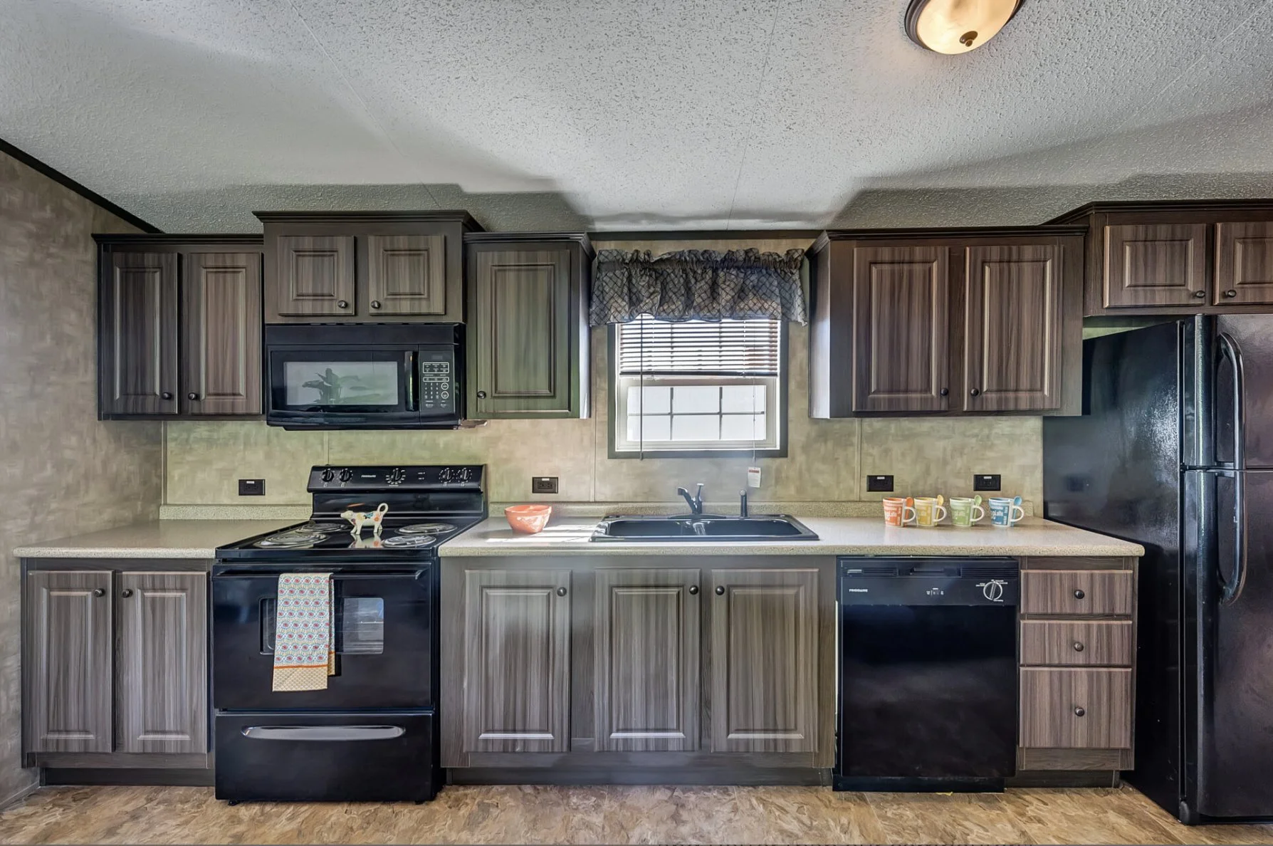 Kitchen with brown wooden cabinets, black appliances including microwave, stove, and refrigerator, a sink under a window with blinds, and a countertop with colorful coffee mugs.