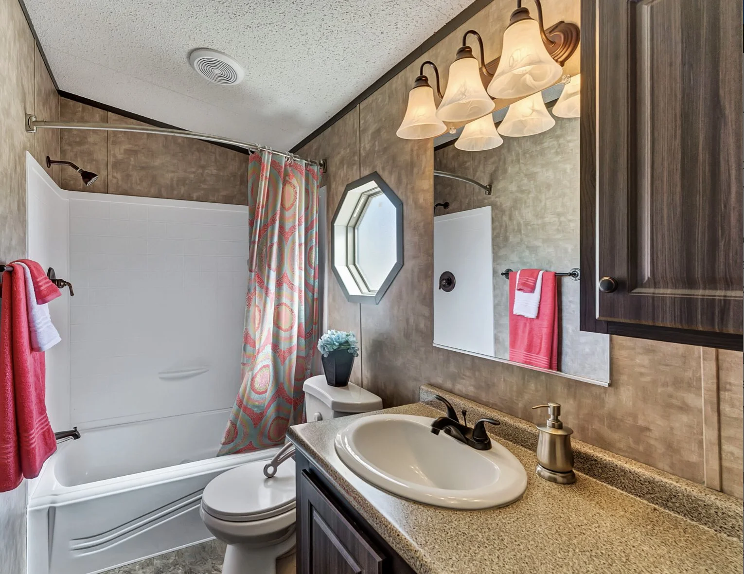 Bathroom with a bathtub, shower curtain, sink with a dark wood cabinet, mirror, wall-mounted light fixture, small octagonal window, and red and white towels.