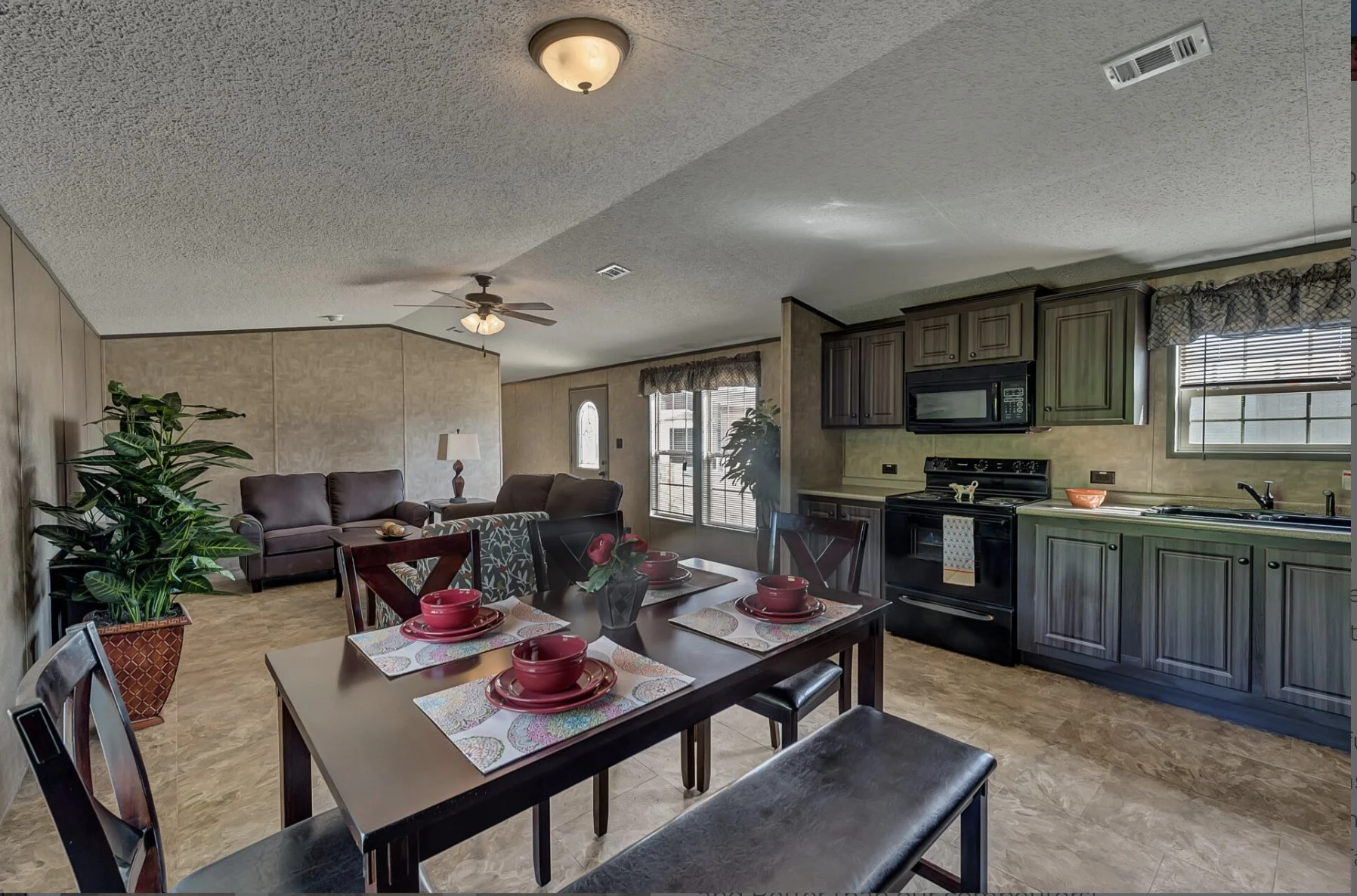 Open-concept living and dining area with a kitchen, featuring a dining table set with red dishware, a dark purple sofa, ceiling fan, large potted plant, and windows with blinds and valances.
