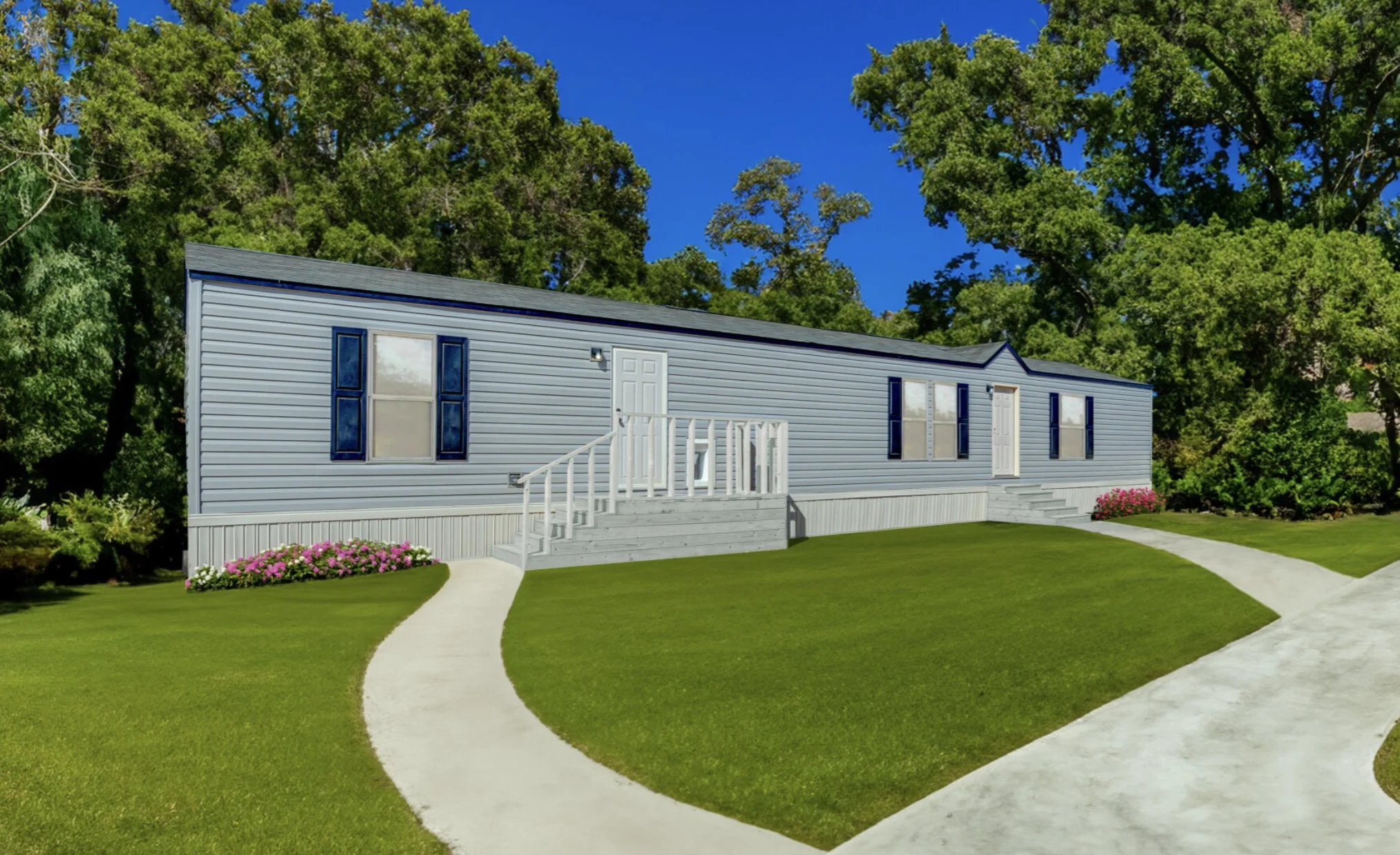 A manufactured home on a grassy lot with a curved concrete walkway leading to the entrances, surrounded by trees and flower beds.