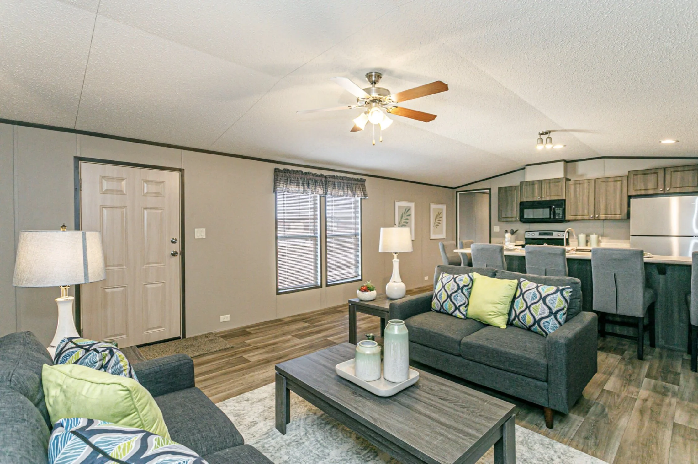Living room with gray couches, colorful pillows, wooden coffee table, and open kitchen with bar stools, gray cabinets, and stainless steel appliances.