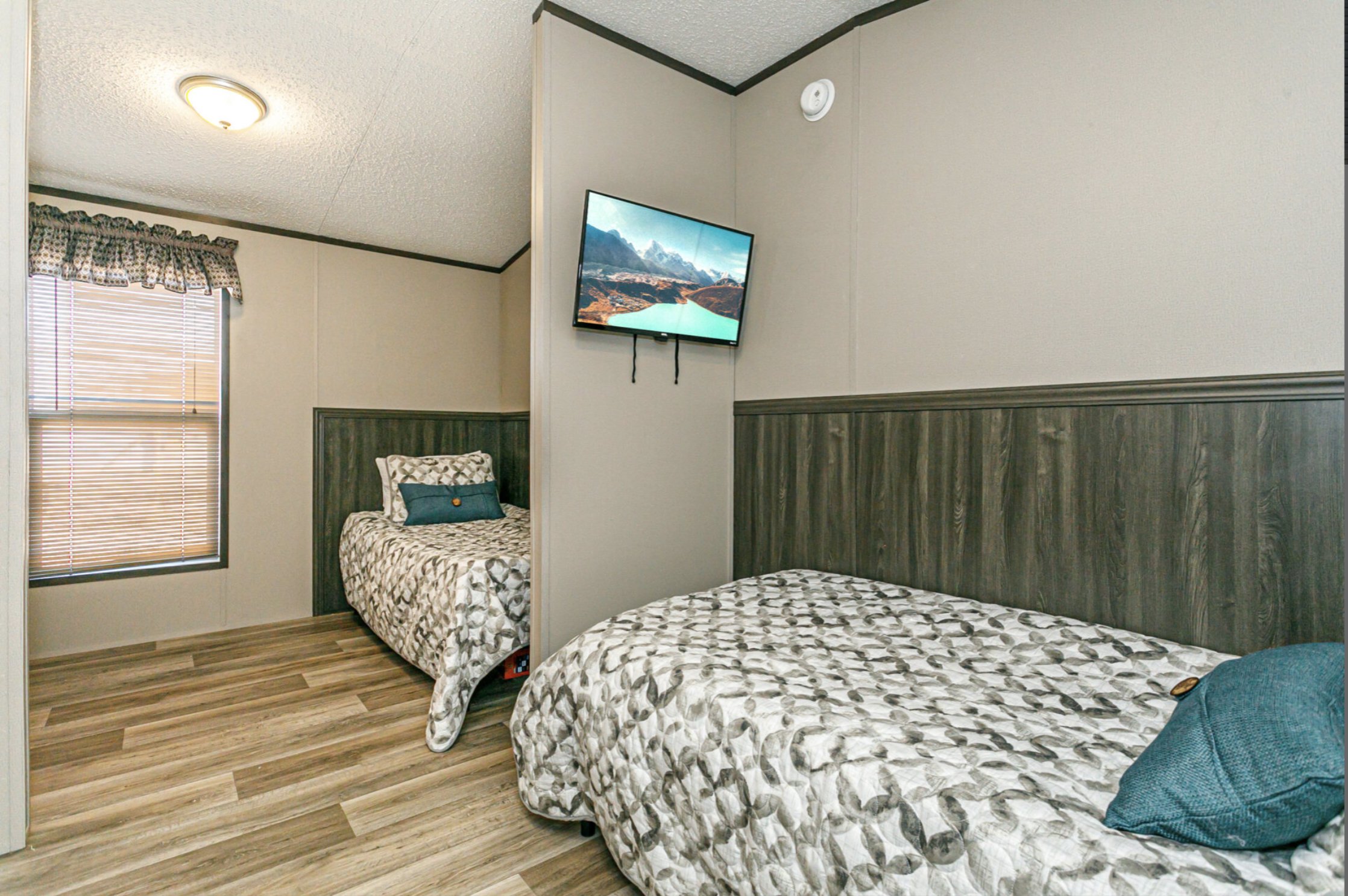 A motel room with two beds featuring matching patterned bedspreads, a window with wooden blinds and a valance, wood paneling headboards, a wall-mounted TV, and a teal pillow on one bed.
