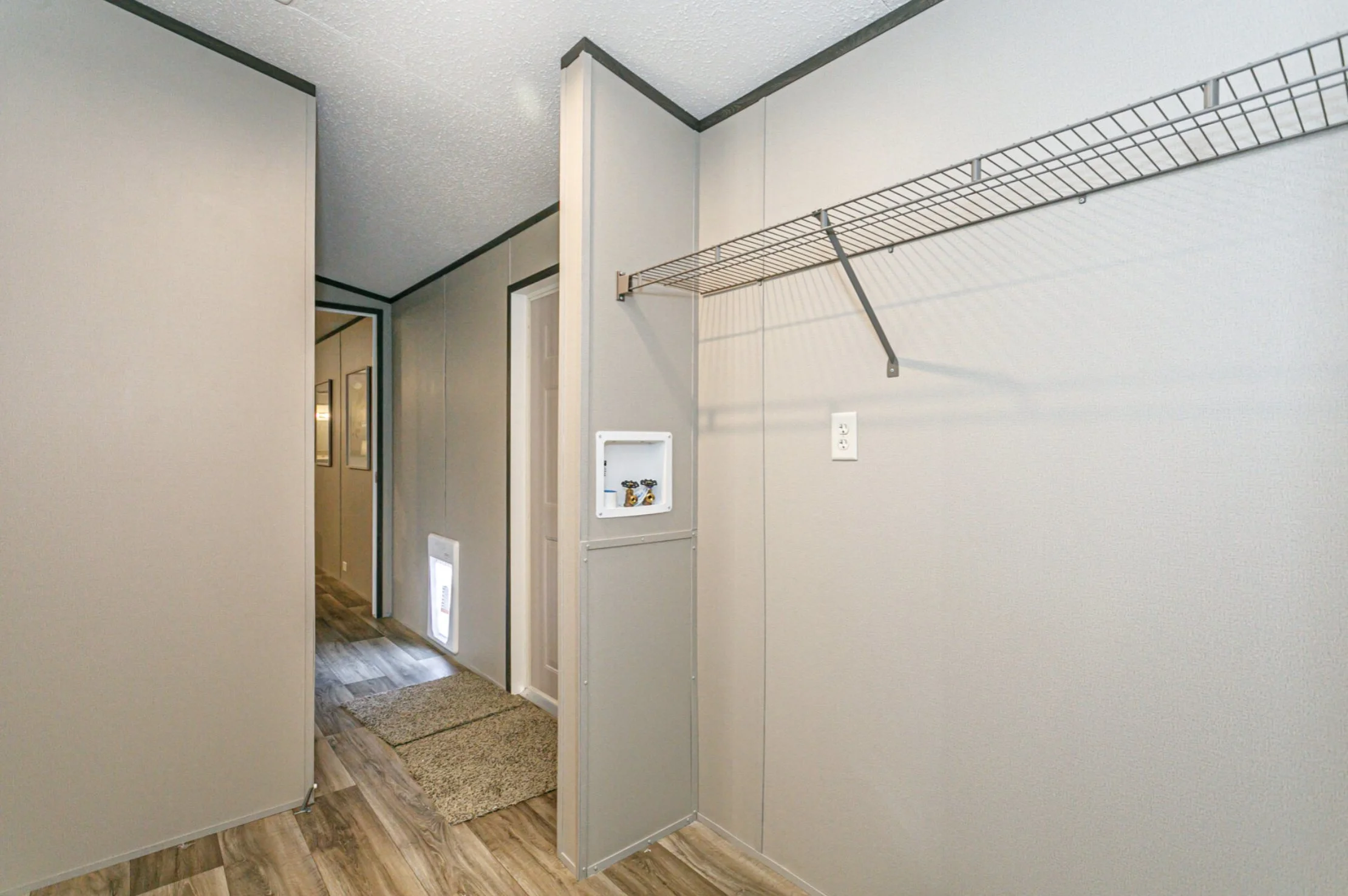 Empty laundry or utility room with a wire shelf on the wall, a small recessed box with decorative items, and a power outlet. An open doorway leads to a hallway with framed pictures and a small grate near the floor.