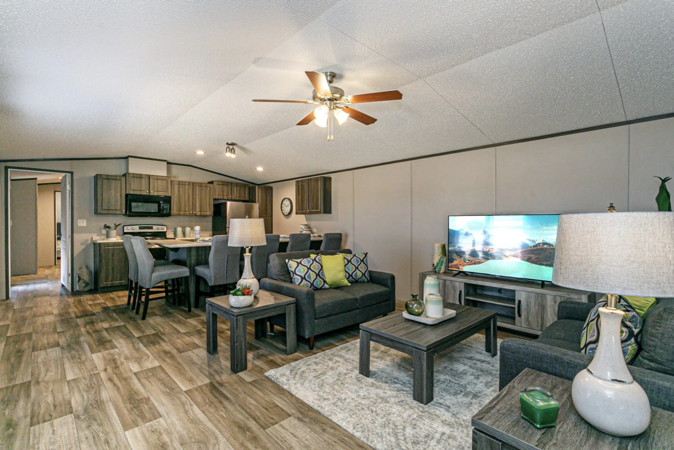 Living room and kitchen area with grey sofas, wooden tables, TV, and kitchen cabinets.
