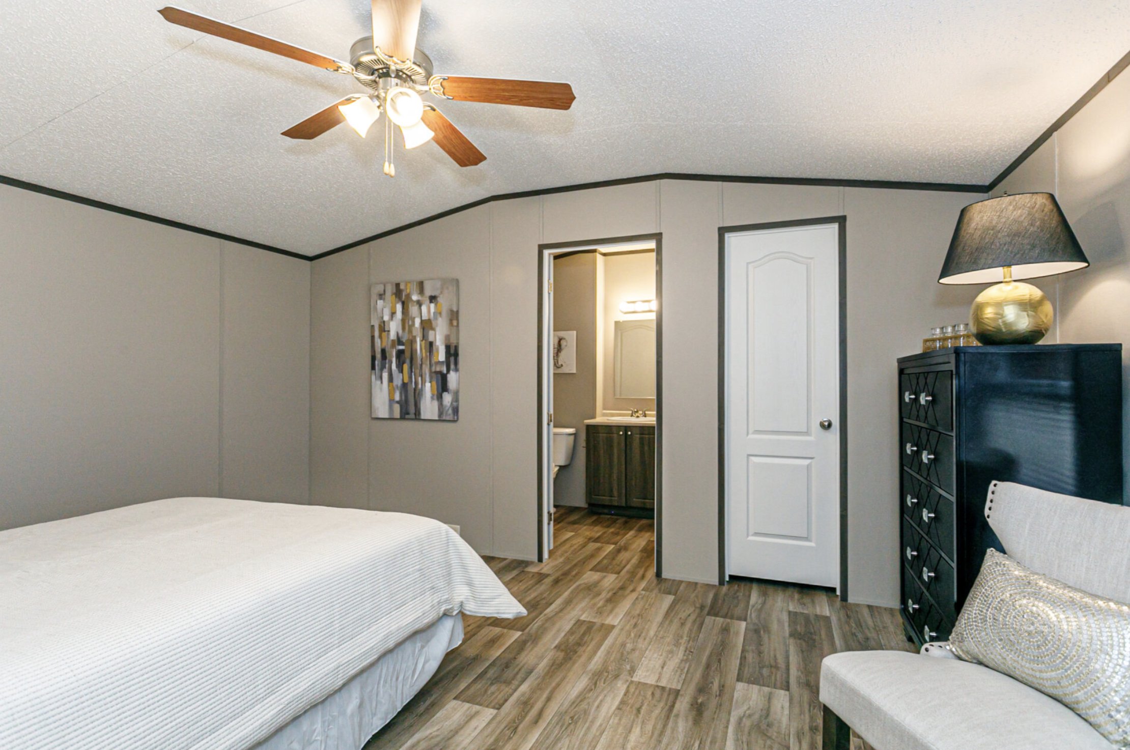 A bedroom with a bed on the left, a black dresser with a gold lamp on the right, a white chair with a decorative pillow, and an open door leading to a bathroom with a vanity and mirror.