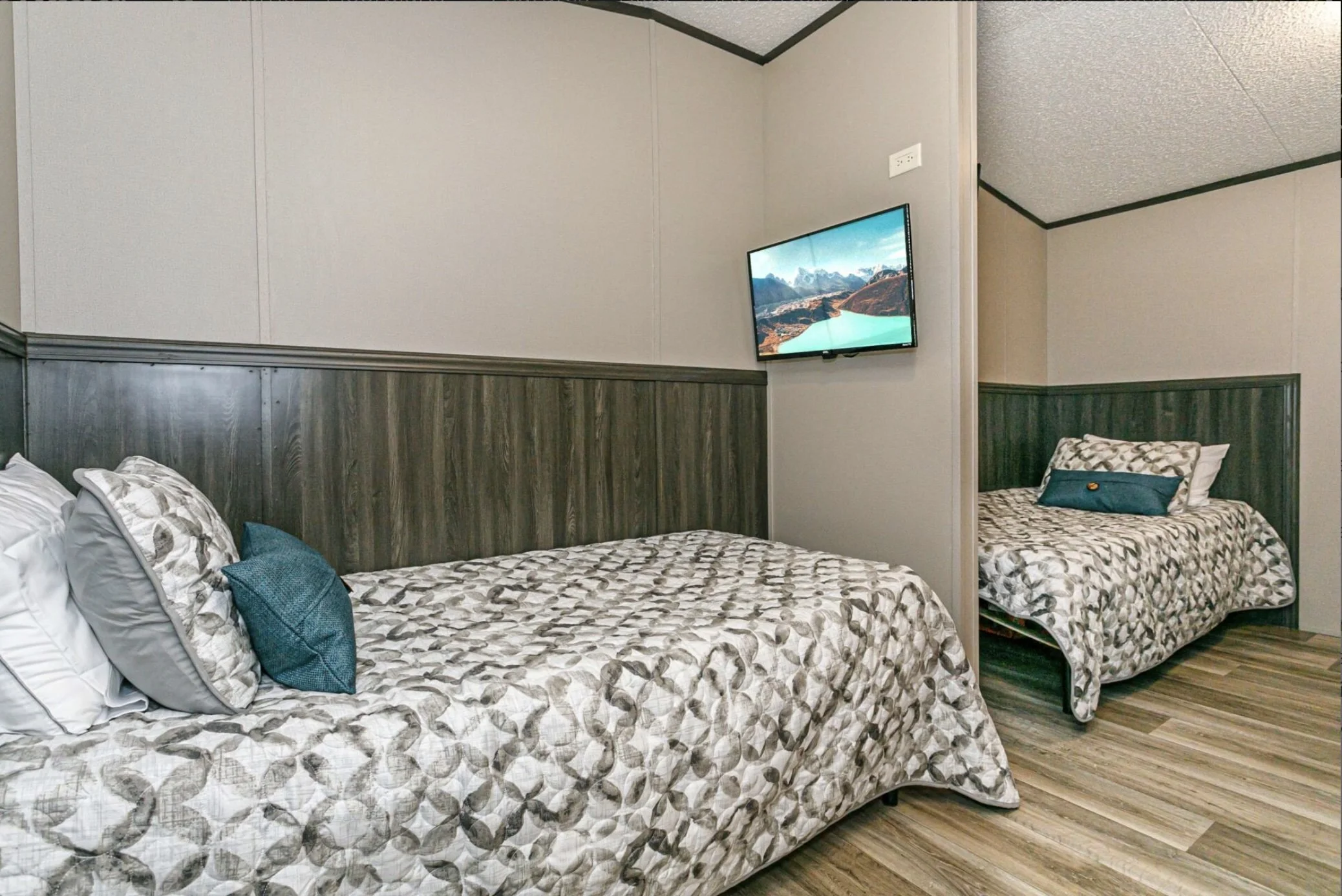 A bedroom with two beds, one larger and one smaller, both with patterned bedding and pillows. A flat-screen TV is mounted on the wall. The room has wood-paneled wall accents and wood flooring.