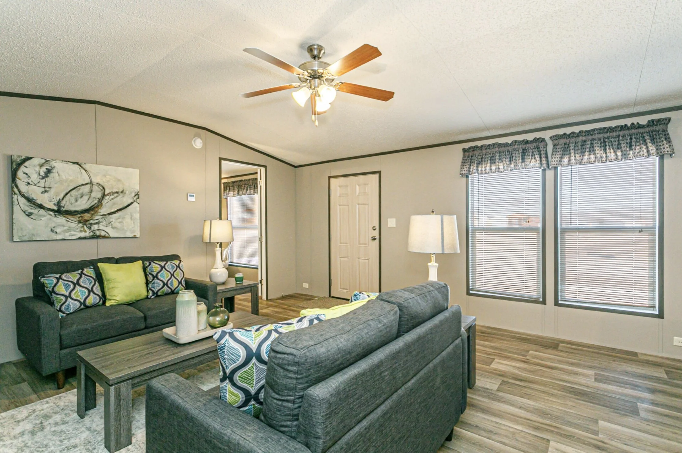 Living room with gray sofas, patterned pillows, a wooden coffee table, table lamps, a ceiling fan with wooden blades, and windows with checkered valances.