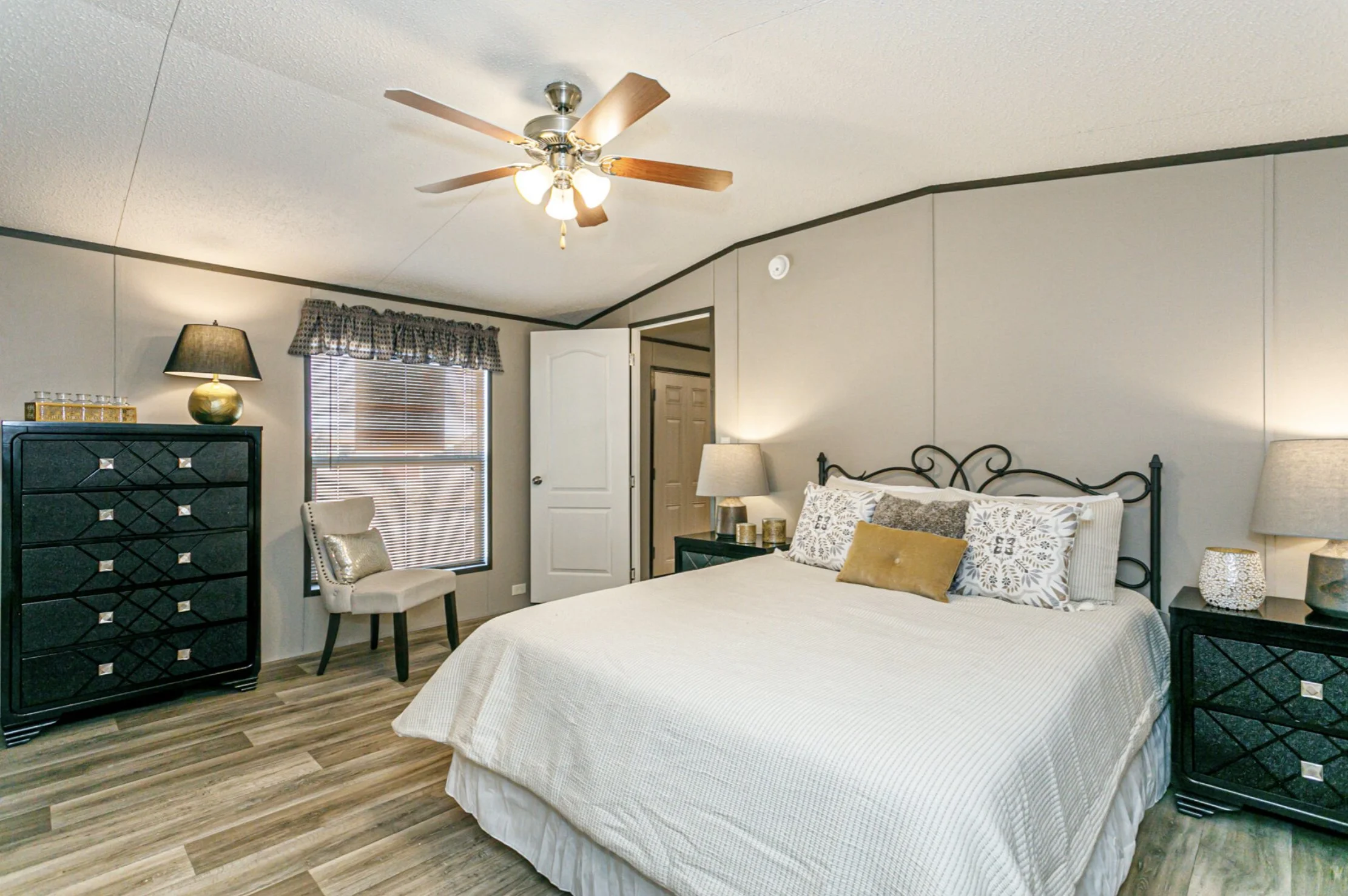 Bedroom with a large bed, black nightstands with lamps, a black dresser with a lamp, a window with blinds and a patterned valance, and a ceiling fan with wooden blades.