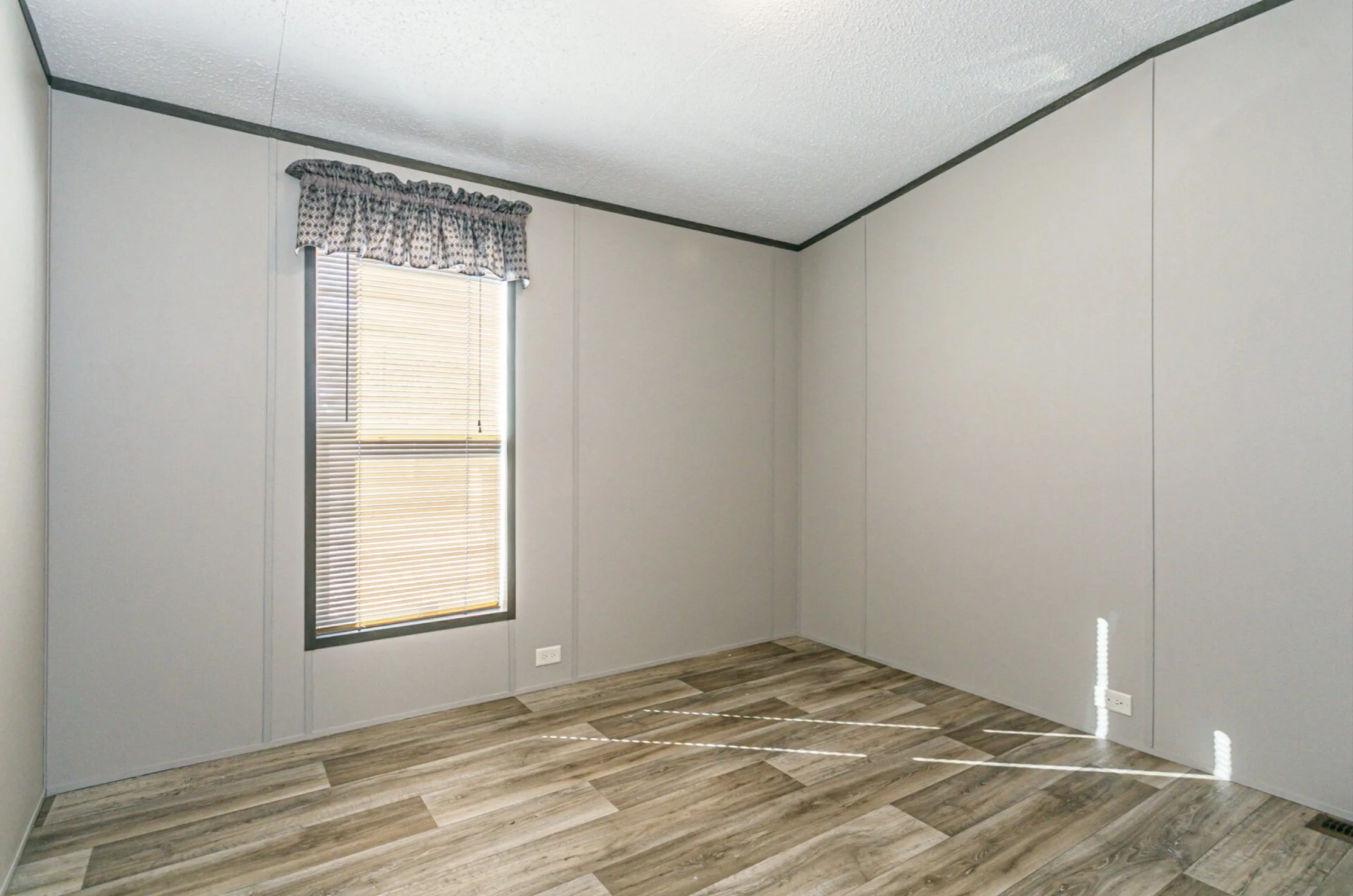 Empty room with a window, decorated with blinds and a valance curtain, wooden floor, and plain light-colored walls.