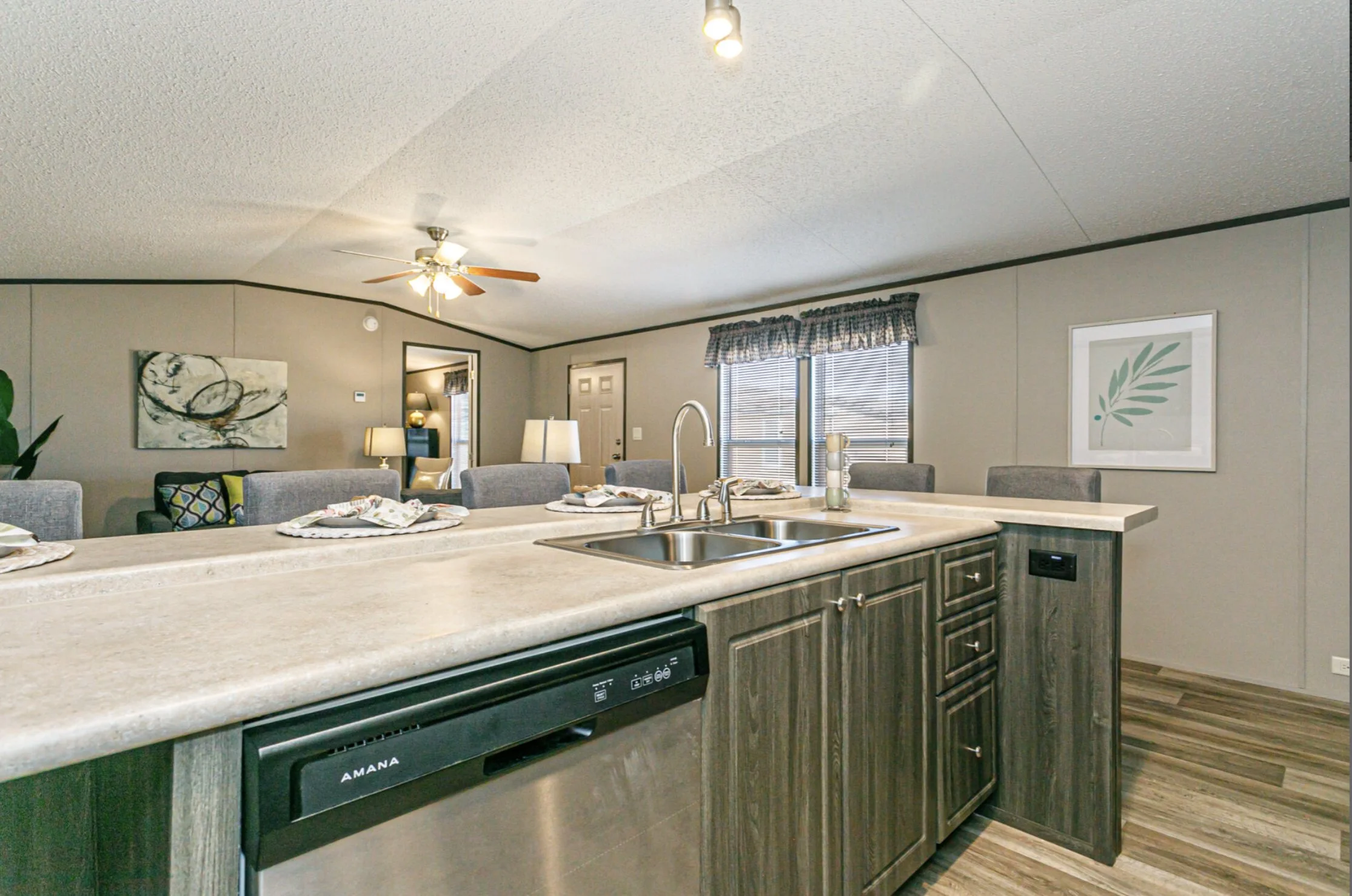 Kitchen with beige countertop, wooden cabinets, stainless steel sink, and open floor plan connecting to living and dining area with gray chairs, wall art, and window with valance
