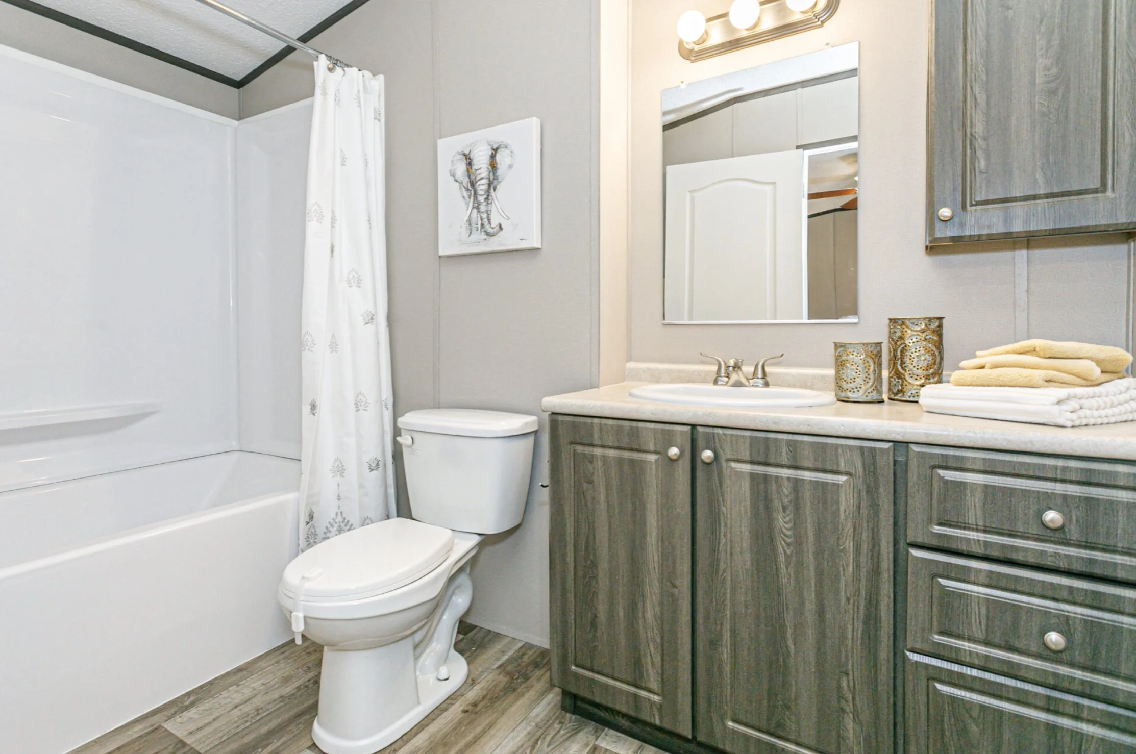 Bathroom with bathtub, toilet, gray cabinets, beige countertop, mirror, decorative candles, and folded towels.