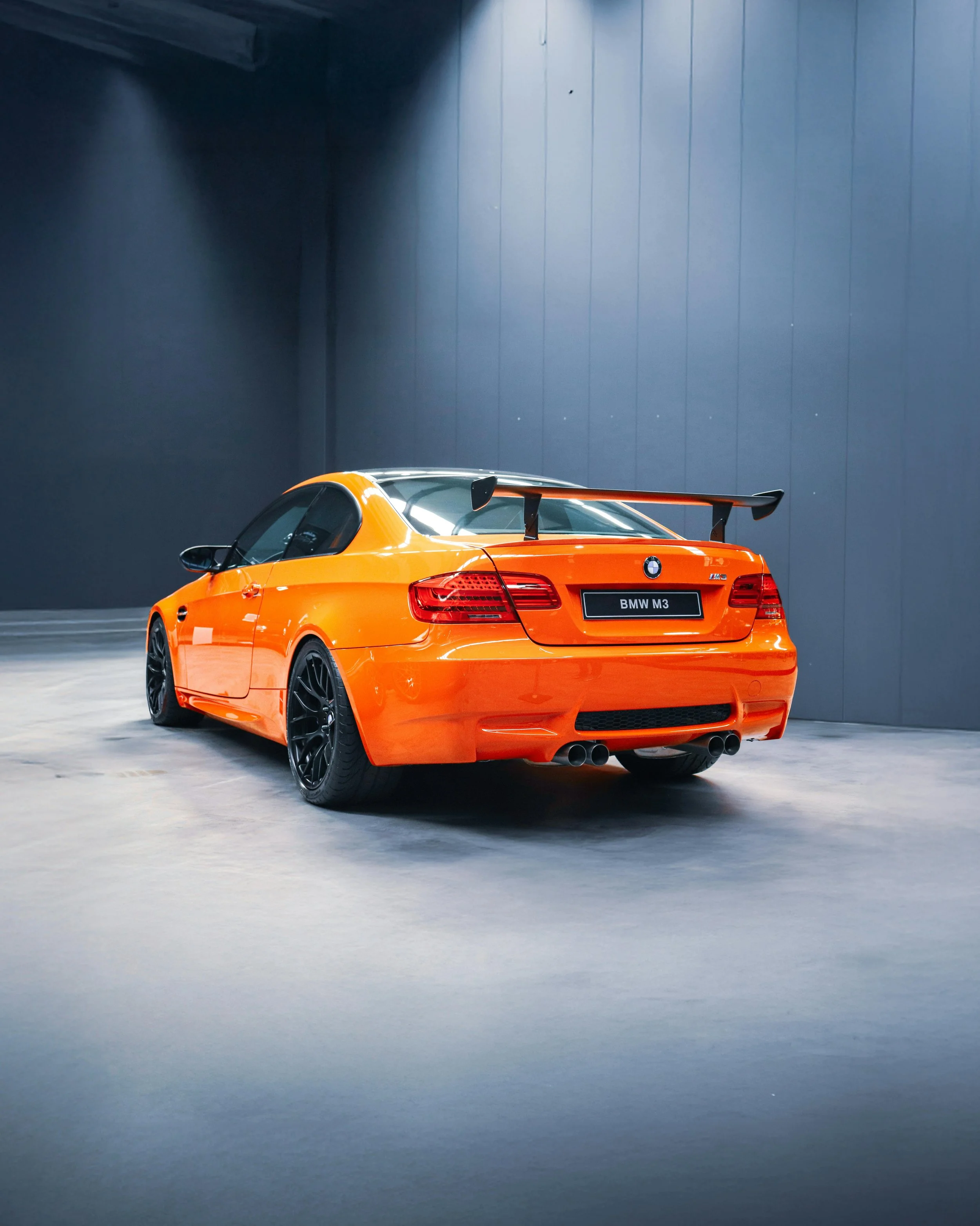 An orange BMW M3 sports car with a large rear spoiler, black wheels, and quad exhausts, parked indoors against a dark wall.