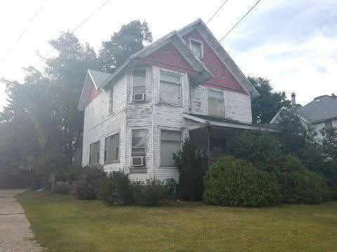 Old, weathered two-story house with peeling paint, surrounded by bushes and trees, under a cloudy sky.