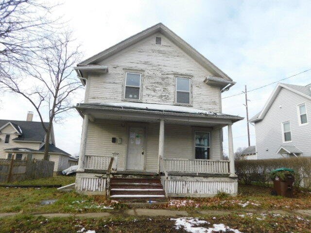 Front view of an old, weathered two-story house with peeling paint, a small porch, and stairs, on a cloudy day with patches of snow on the yard.