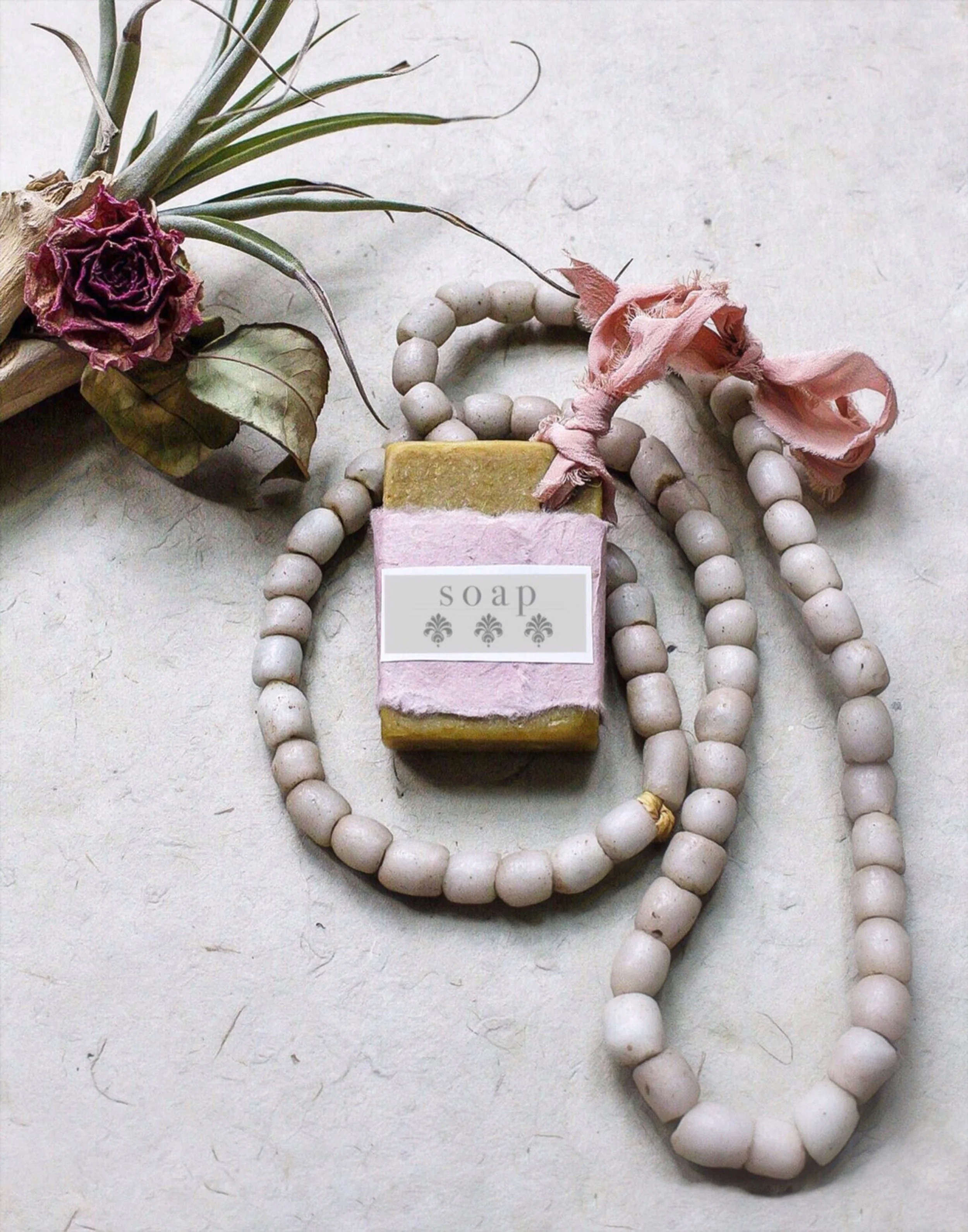 A handmade soap bar with pink and yellow layers, labeled 'soap' with fleur-de-lis symbols, surrounded by white bead necklaces, and decorated with dried flowers and a pink ribbon.