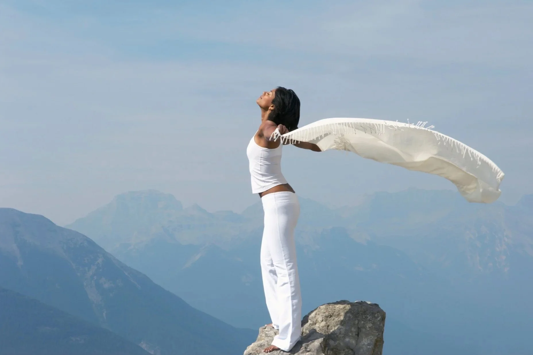 Woman standing on a mountain rock with arms open to the sky and a scarf flowing in the wind.