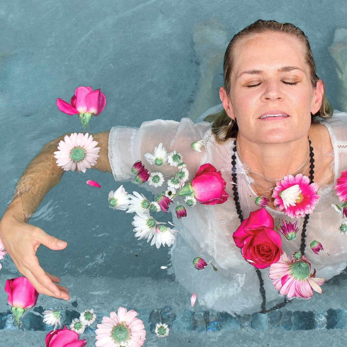 A woman resting in blue muted water with brightly colored hot pink and white flowers floating on top of the water. She is wearing a sheer white top and a strand of male beads. Leslie Storms