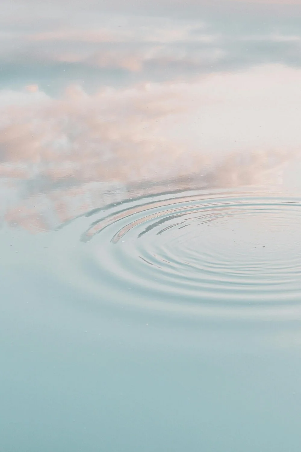 Calm water surface with gentle ripples, reflecting soft pastel-colored sky with clouds.