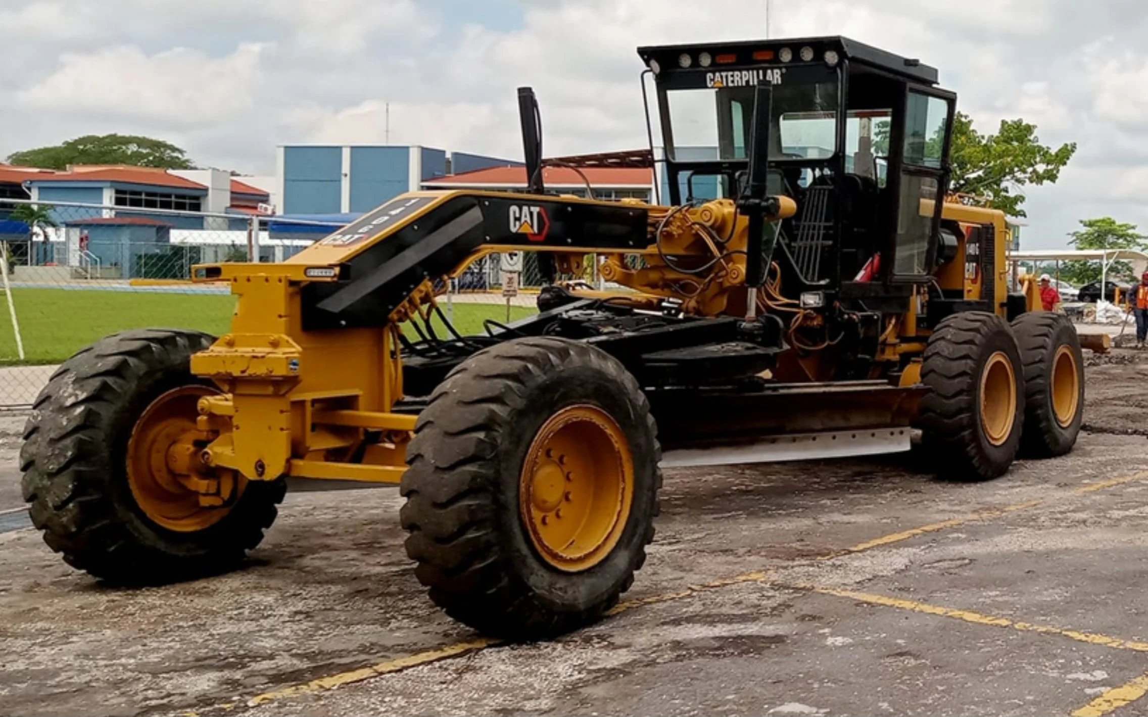 A large yellow Caterpillar roller compactor parked outdoors on a construction site with a building and trees in the background.