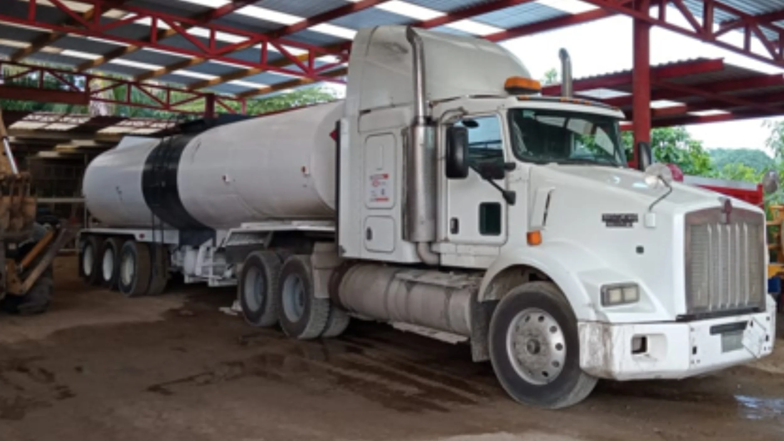White tanker truck parked inside a shed with a dirt floor, red metal roofing, and some construction equipment nearby.