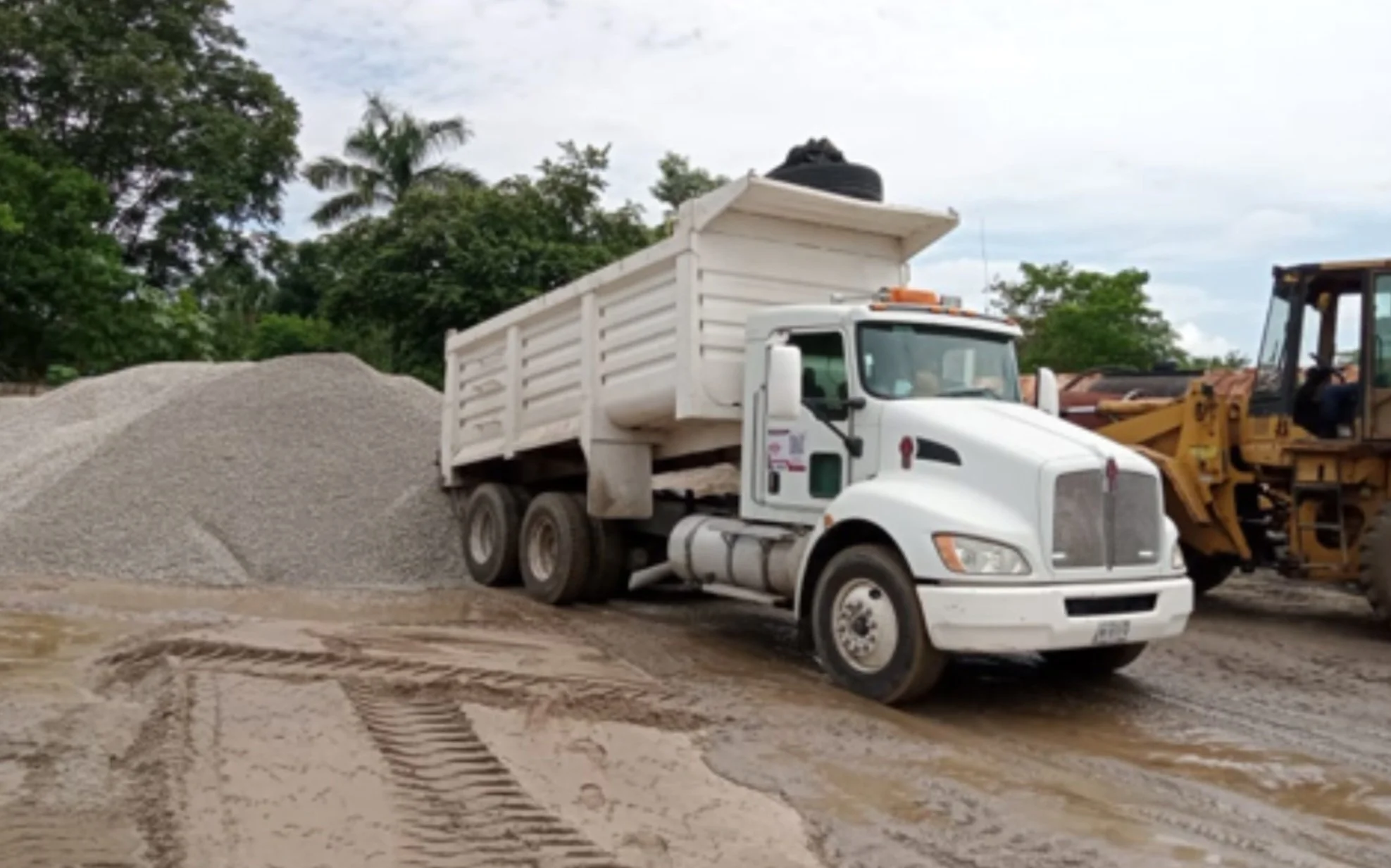 A white dump truck on a construction site, with a large pile of gravel behind it and a yellow bulldozer on the right. The ground appears muddy with tire tracks.