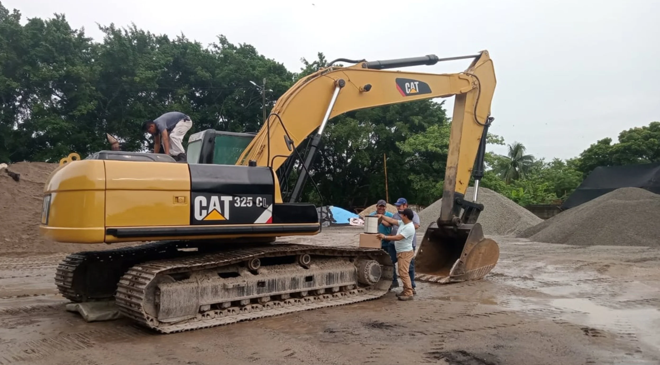 People standing next to a yellow Caterpillar 325C excavator on a construction site with piles of gravel and dirt, trees, and overcast sky in the background.