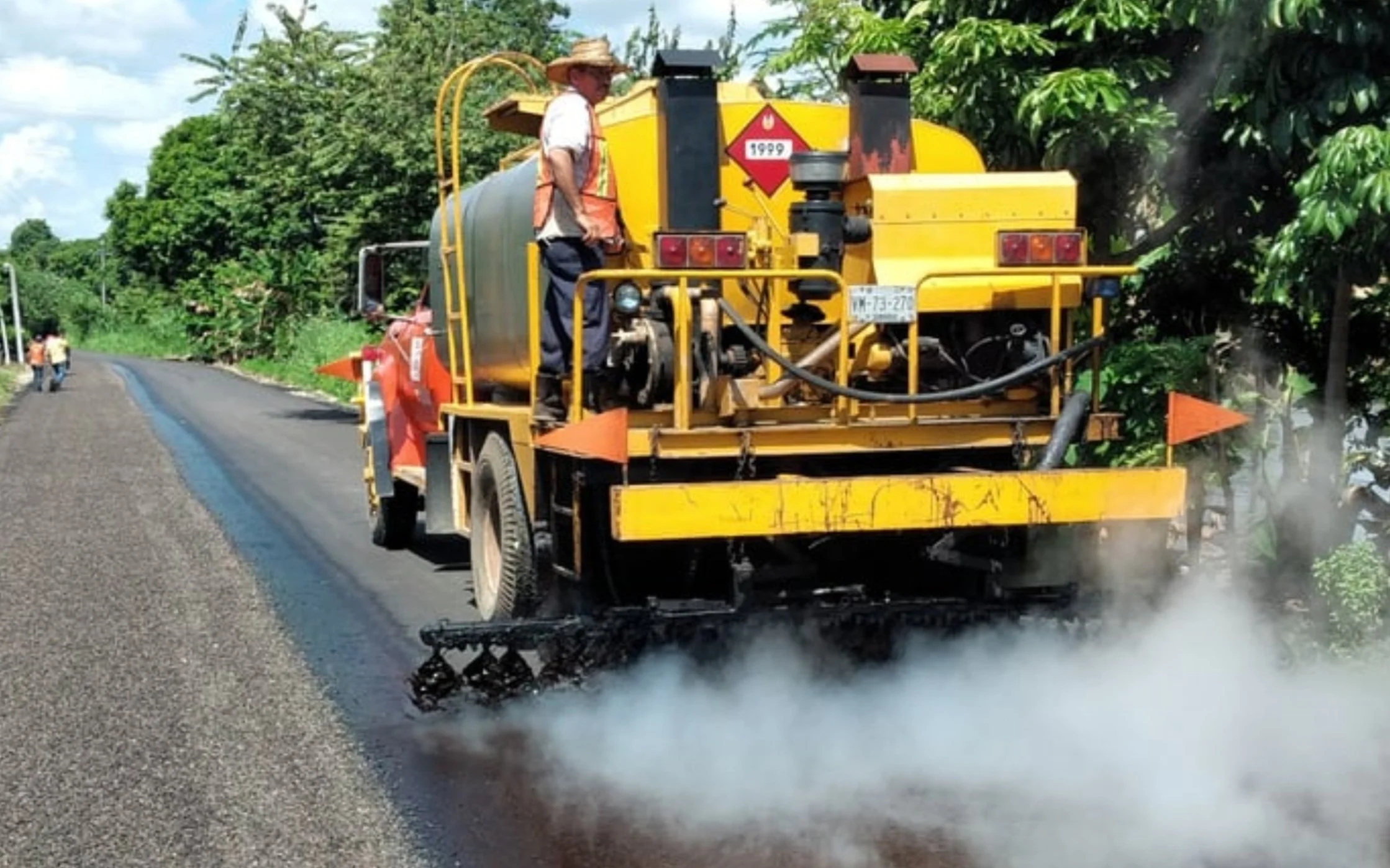 A yellow road construction machine sprays hot asphalt onto a road surface, with a worker standing on top, surrounded by green trees and a partly cloudy sky.