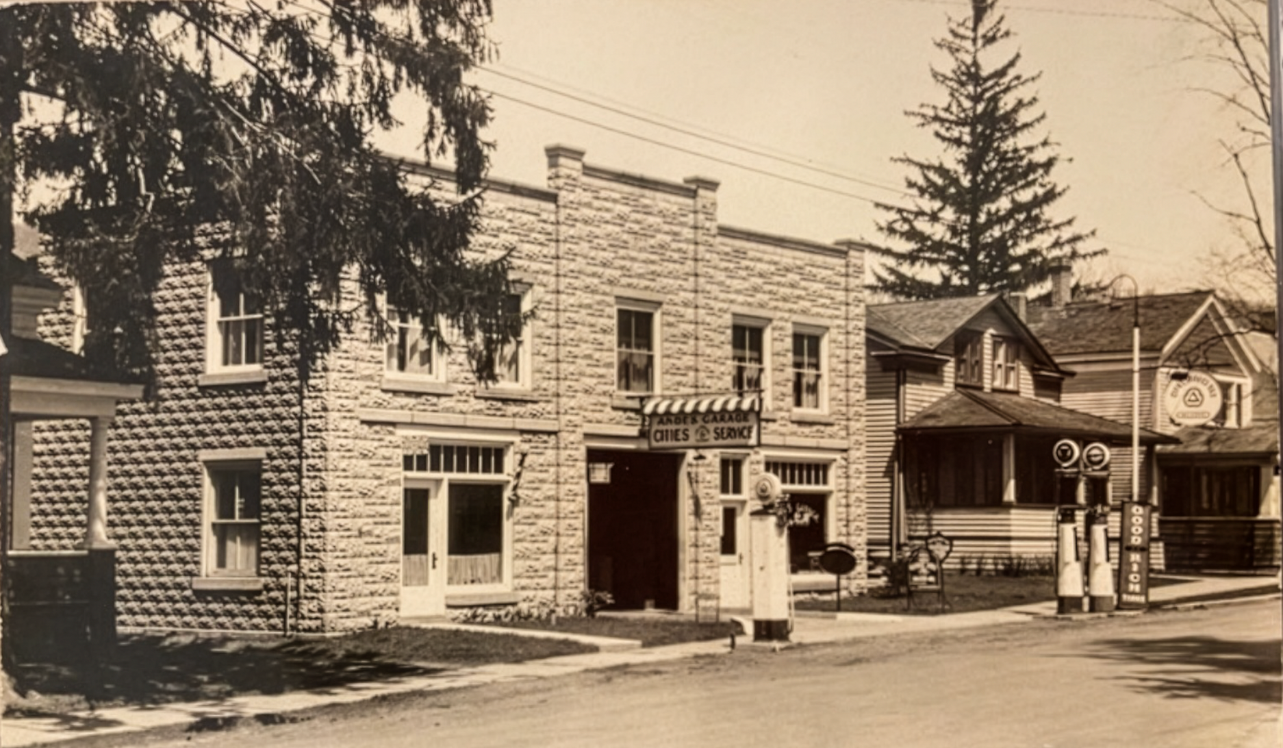 Old black-and-white photo of a commercial building with a sign that reads "Andes Garage, Gines Service," a small lawn, a sidewalk, and two vintage gas pumps in front.