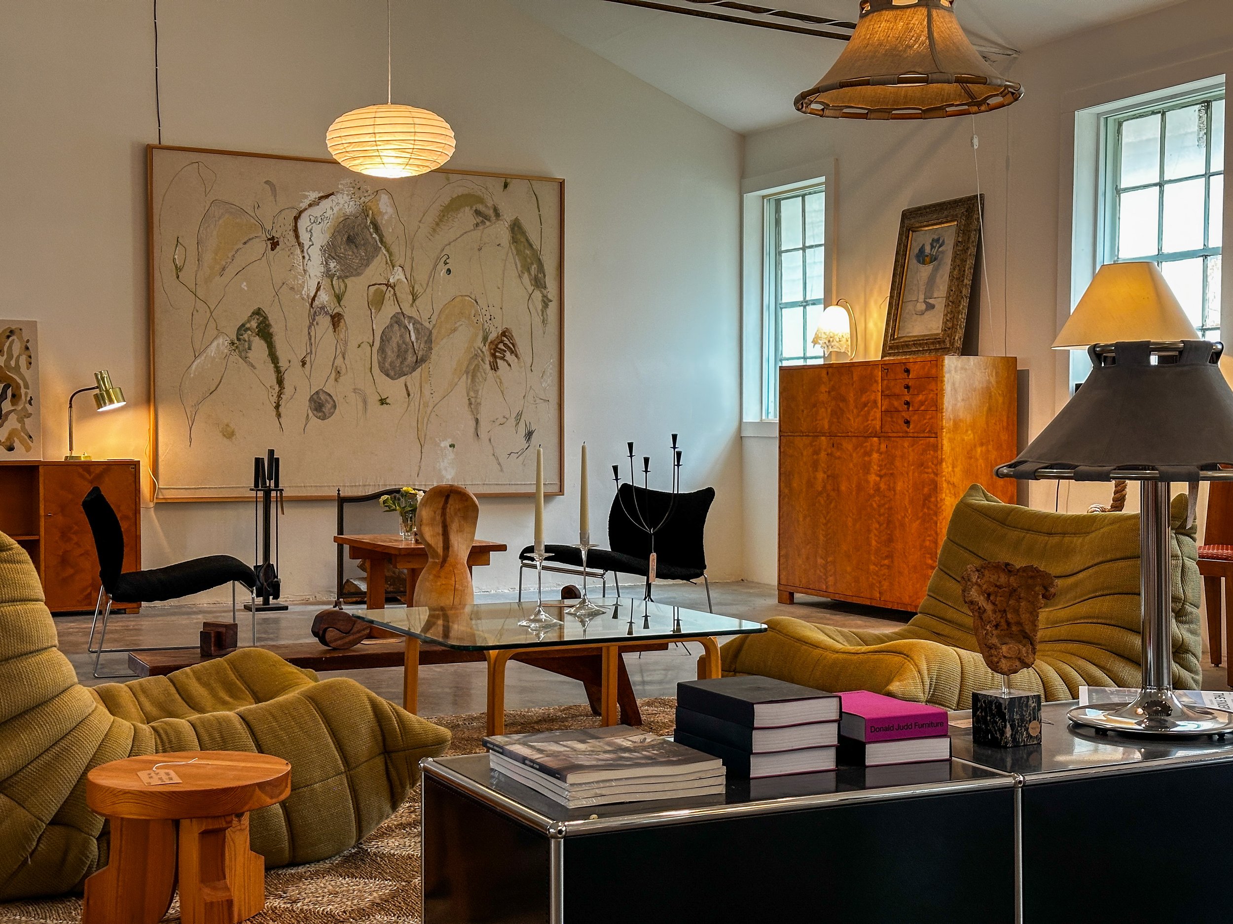 Interior of a living room with mid-century modern furniture, abstract artwork, and various lamps, some turned on, with large windows letting in natural light.