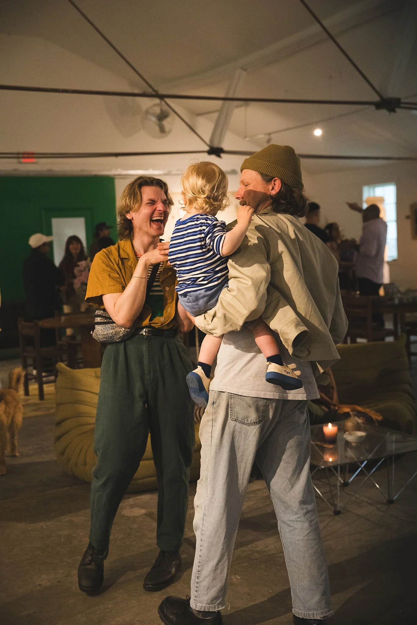 A woman laughing and a man holding a young child in a striped shirt and blue shoes in an indoor setting with other people in the background.