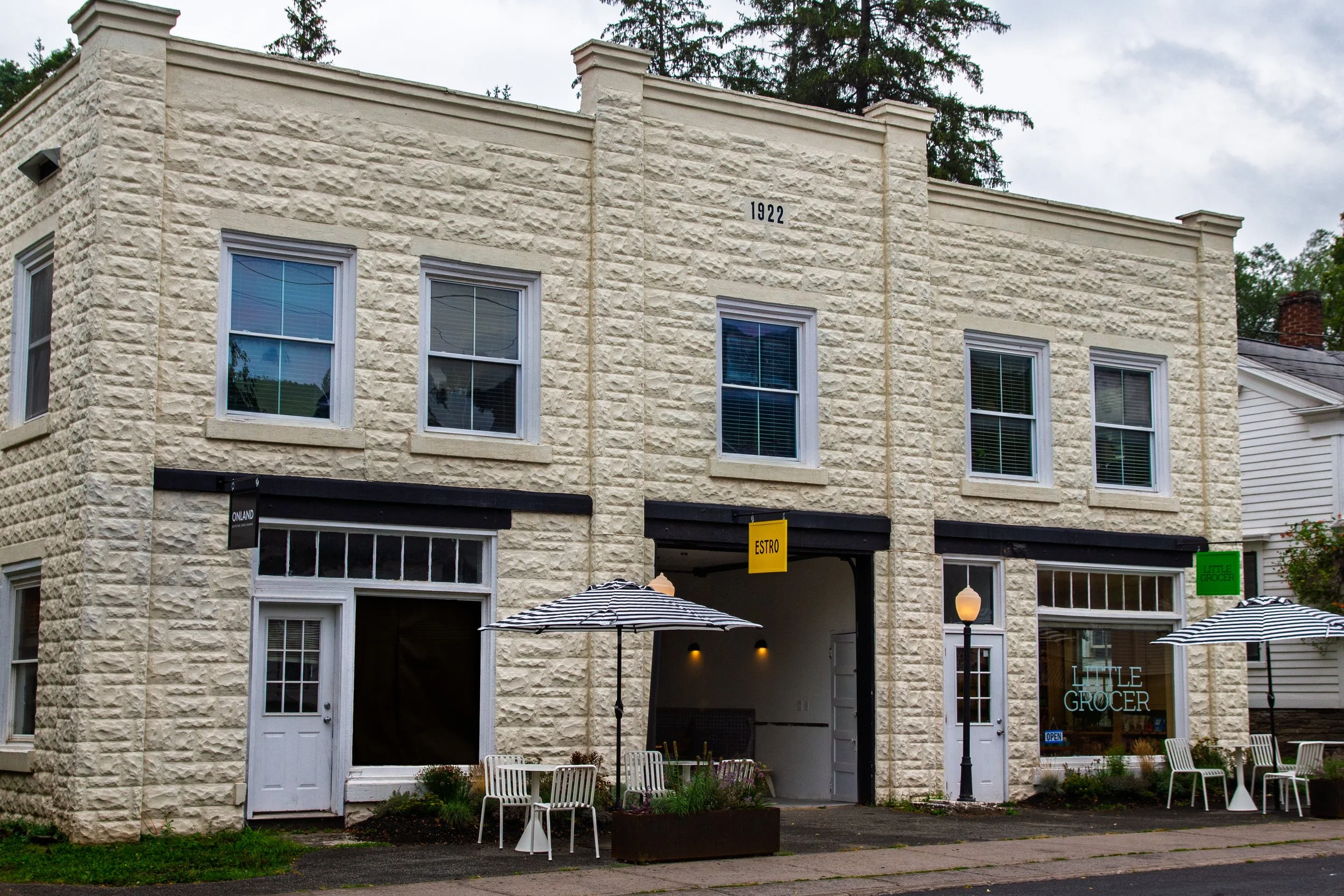 A two-story building with a stone exterior, multiple windows, and storefronts on the ground level. There are outdoor tables with umbrellas, and signs that read 'LITTLE GROCER,' 'ESTRO,' and 'ONLAND.'
