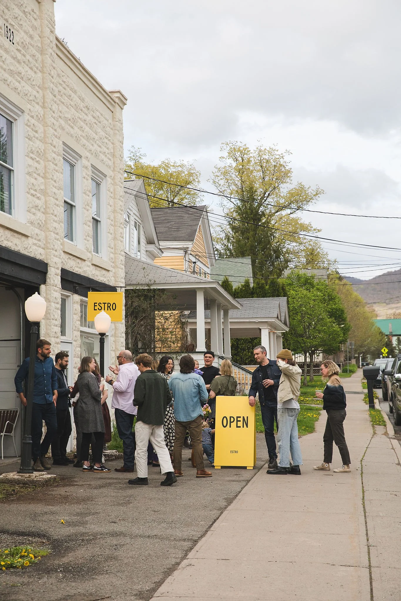 People standing and socializing outside a building with a yellow sign reading 'OPEN' and another sign with 'ESTRO'. The setting appears to be a small town or neighborhood with residential houses and tree-lined streets.