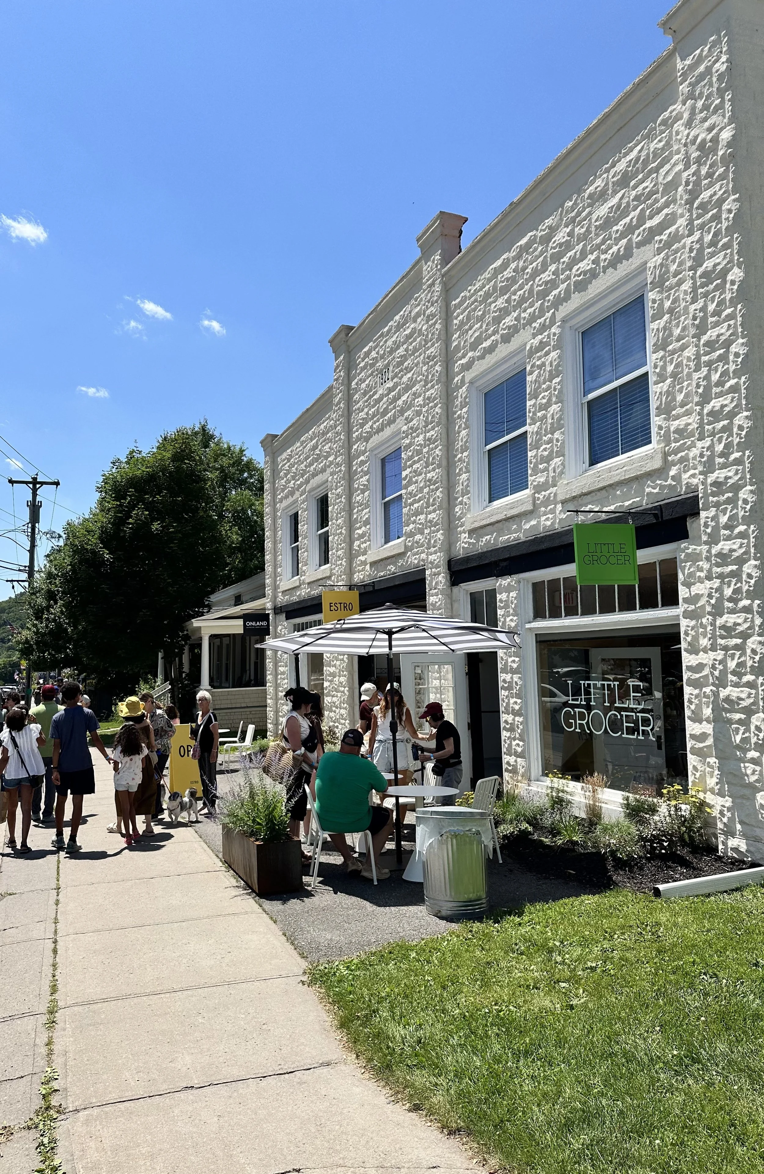 People gather outside a white brick building with a sign for Little Grocer, some sitting under an umbrella, others walking along the sidewalk on a sunny day.