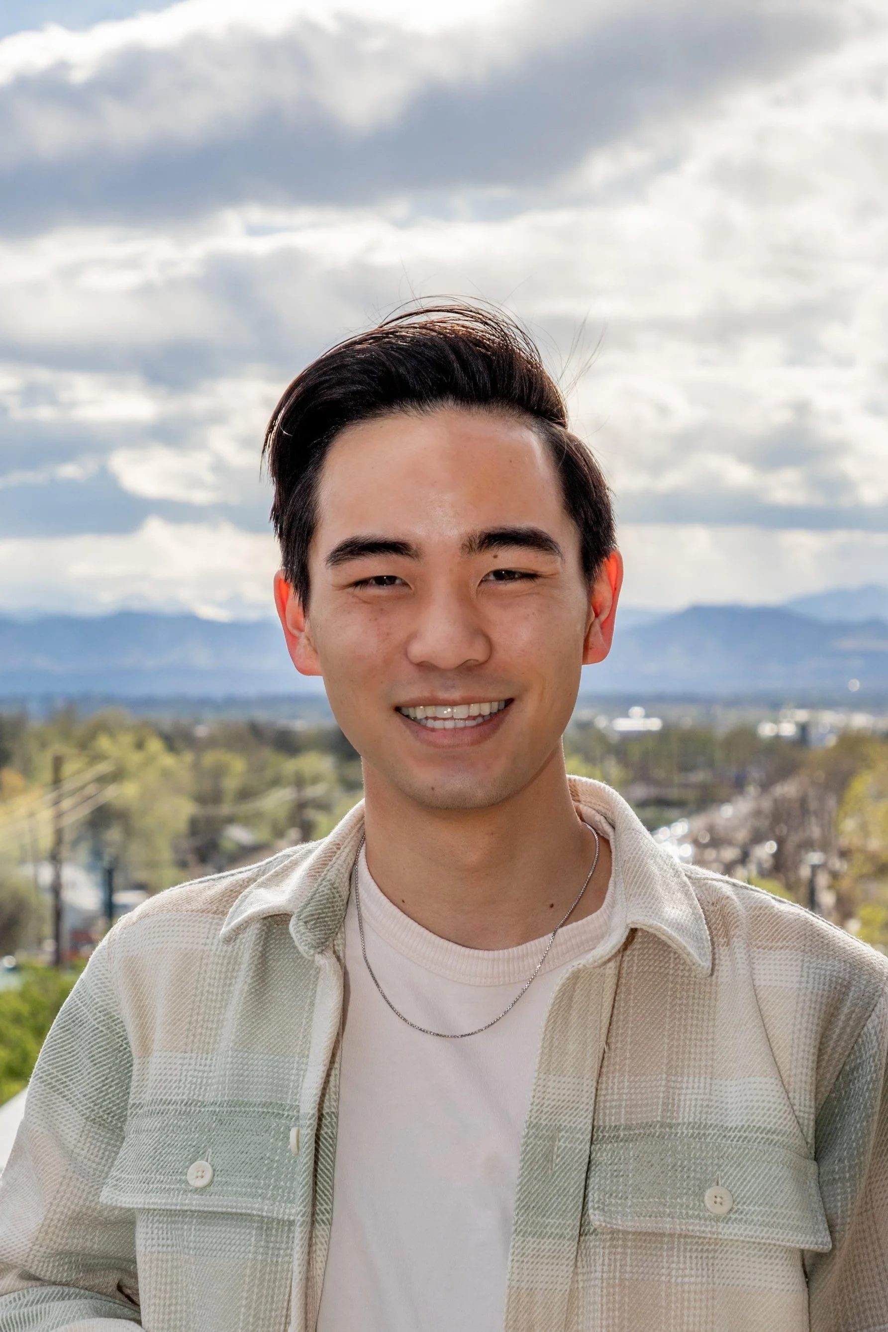 A young man with short, dark hair smiling outdoors with a scenic background of mountains, cloudy sky, and distant trees and buildings.