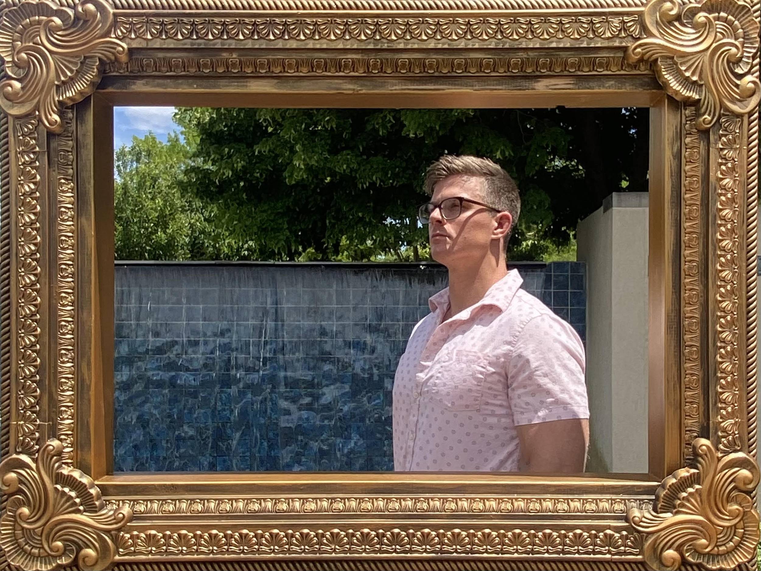 A man with glasses and short hair standing outdoors, looking to the right, seen through an ornate gold-colored picture frame. There is a large blue-tiled water feature and green trees in the background.