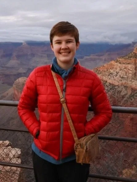 A smiling young person with short brown hair wearing a red jacket and a brown crossbody bag, standing in front of a canyon landscape with layered rock formations and cloudy sky.