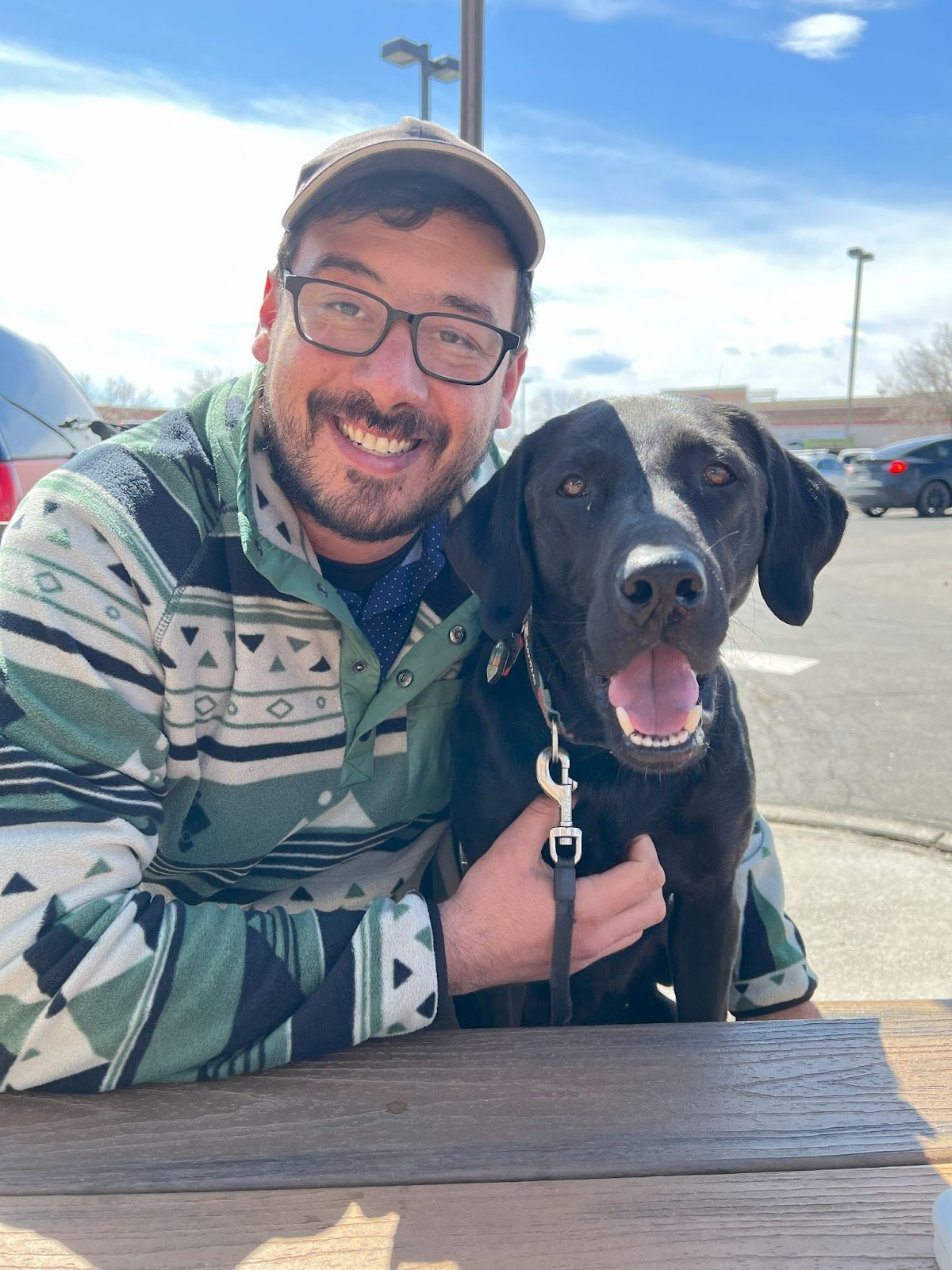 A man with glasses and a beard smiling, wearing a patterned jacket and a tan cap, sitting outside with a black dog with a white chest. They are at a picnic table in a parking lot on a sunny day.