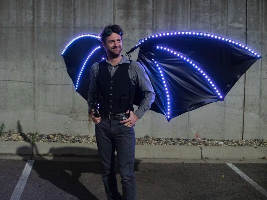 A man standing in a parking lot at night with illuminated bat wings attached to his back.